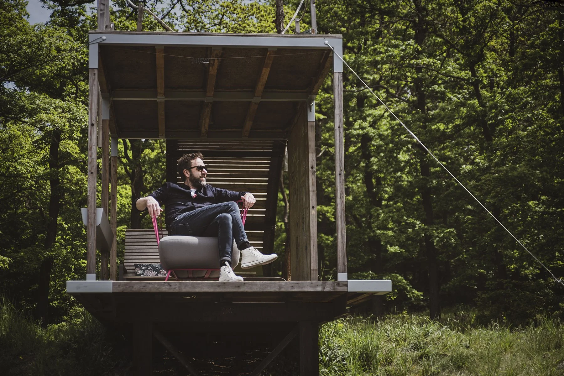 Osoba relaxující v křesle na dřevěné terase uprostřed lesa s výhledem do zeleně, produktová lifestyle fotografie.