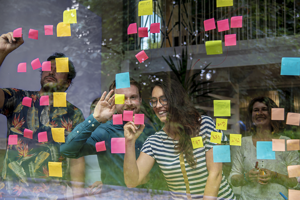 Lidé při týmovém brainstormingu lepící barevné sticky notes na skleněnou stěnu, firemní brand fotografie.