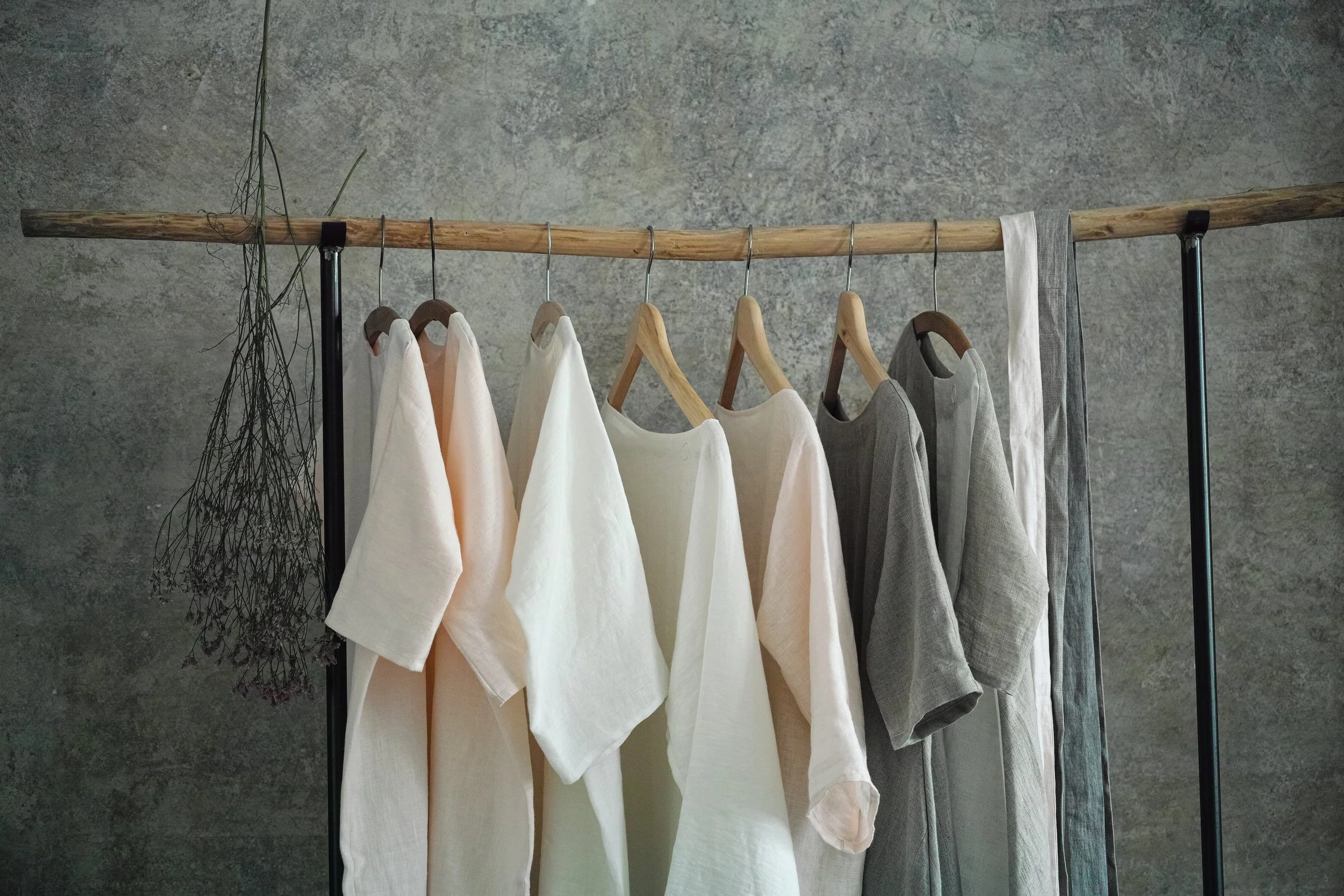 Kolekce lněných a přírodních oděvů Novadiva zavěšených na dřevěné tyči v minimalistickém prostoru.