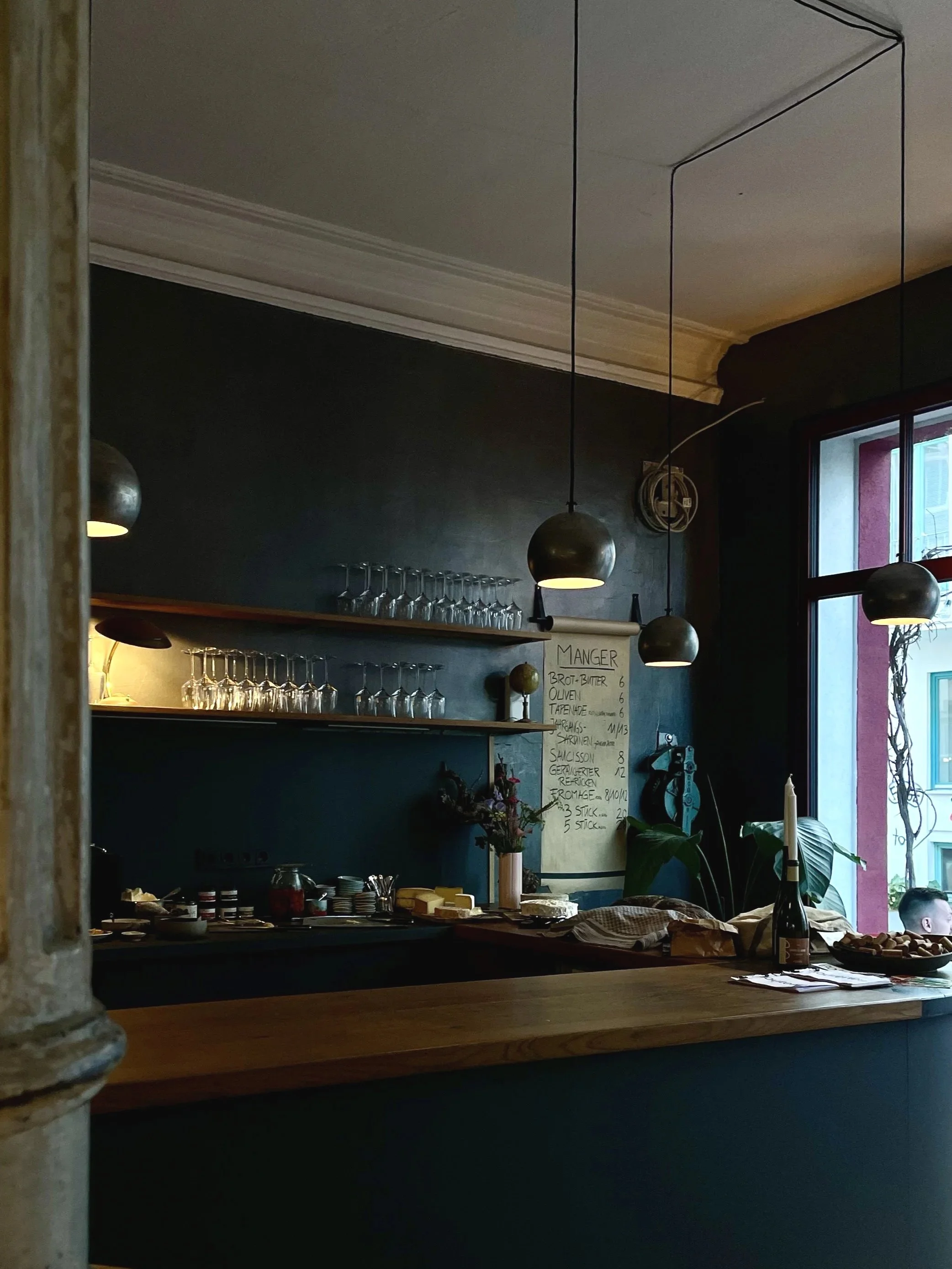 Blick in ein gemütliches Café mit dunklen Wänden, hängenden Lampen, einer Speisekarte an der Wand, einer Fensterfront und einer Essensauslage auf der Theke.