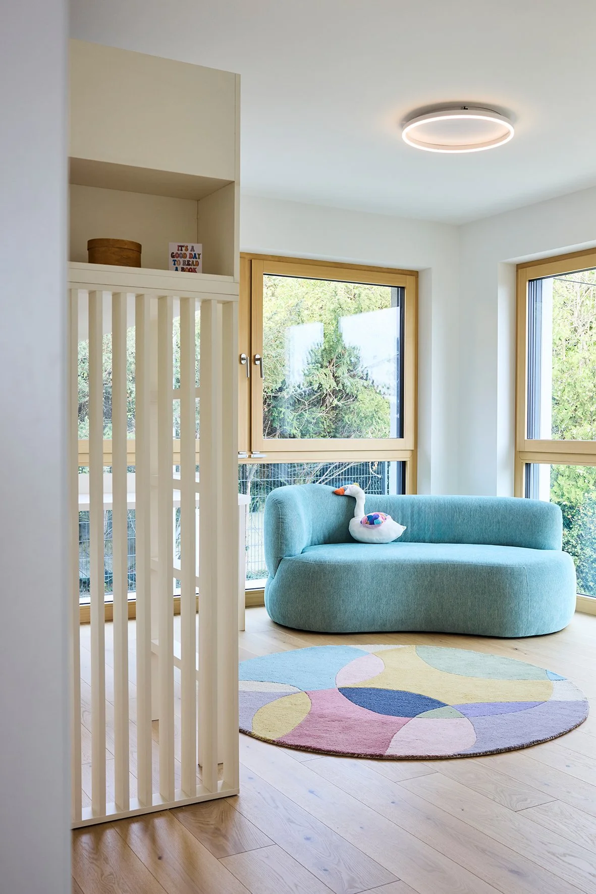 Ein helles, modernes Kinderzimmer mit großen Fenstern, einem hellblauen Sofa, einem bunten runden Teppich mit geometrischen Mustern und einer kleinen weißen Gans- oder Entenfigur auf dem Sofa.
