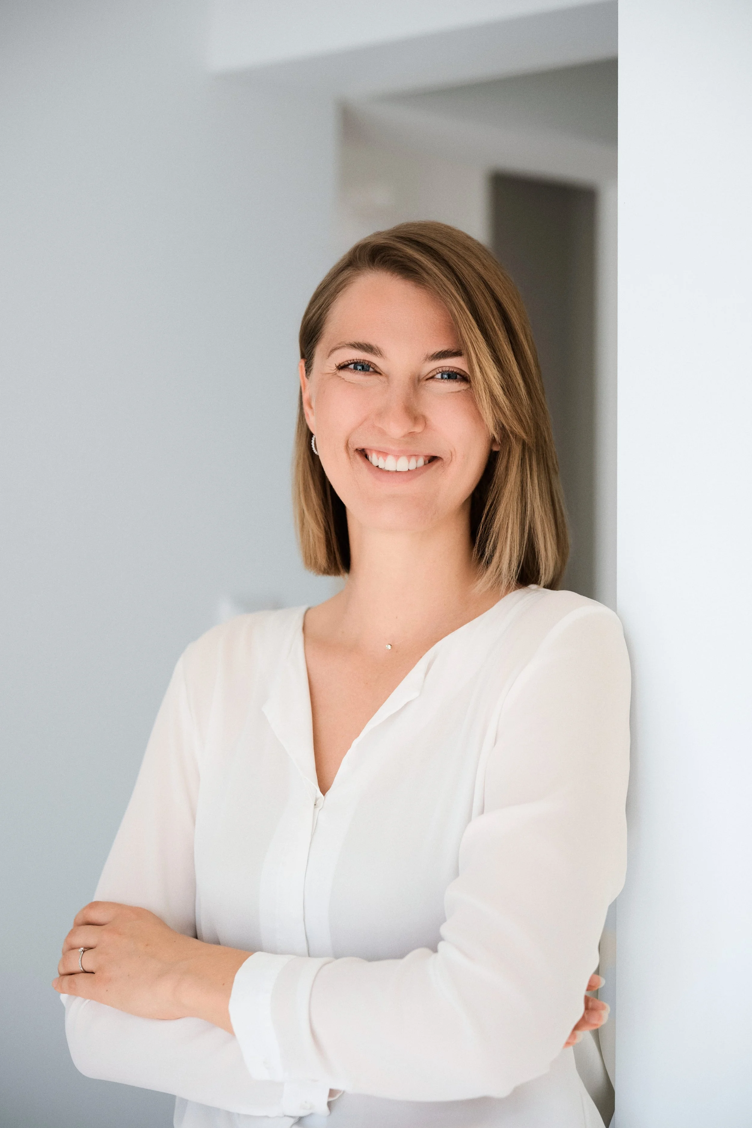 Junge Frau (Vanessa Schaal) mit kurzen braunen Haaren, weißem Hemd, lächelnd, vor weißer Wand in modernem Raum.
