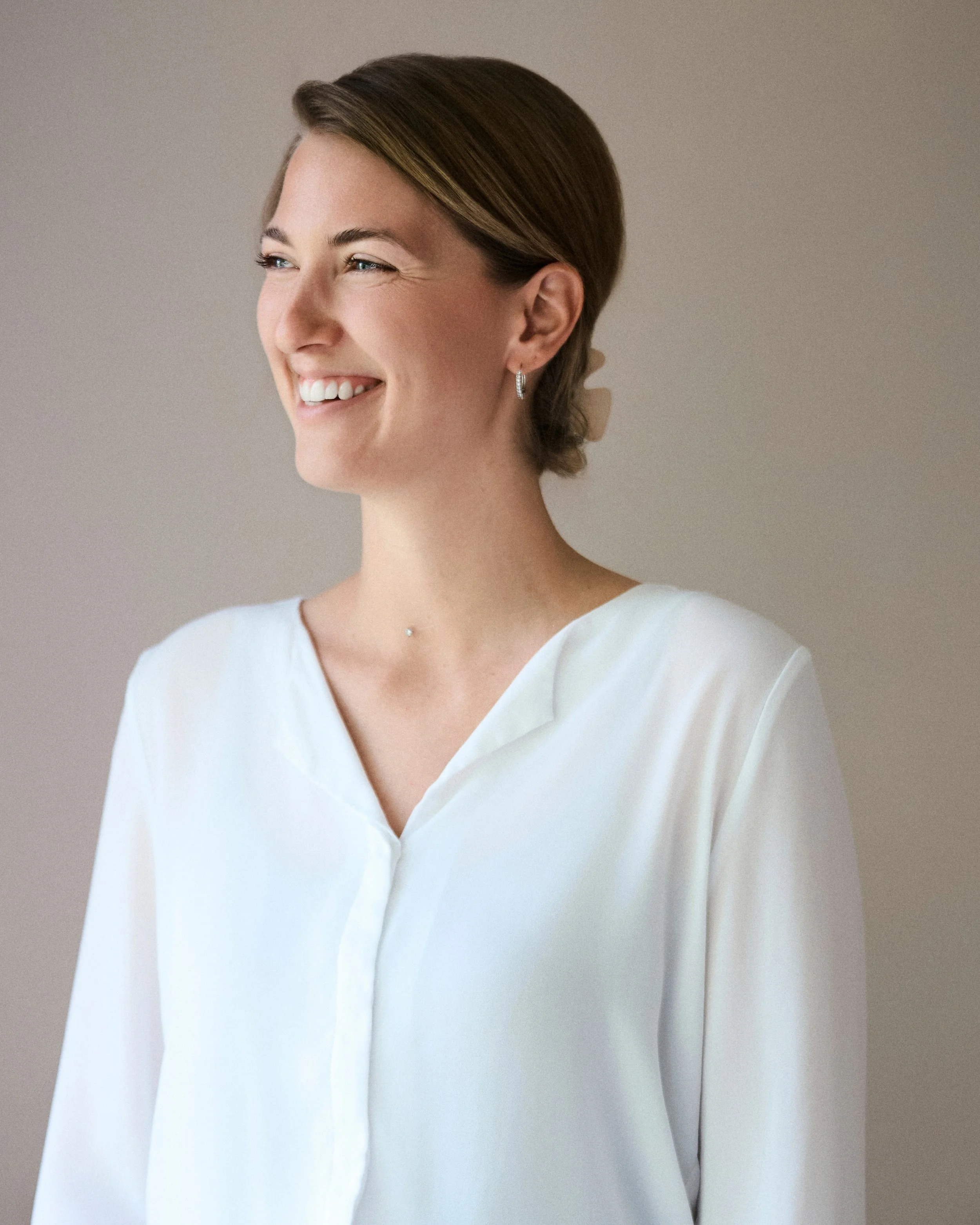 Lächelnde Frau (Vanessa Schaal) in Weißem Oberteil, Seitenansicht, kurze braune Haare, Ohrringe, kleine Halskette, gegen neutralen Hintergrund.