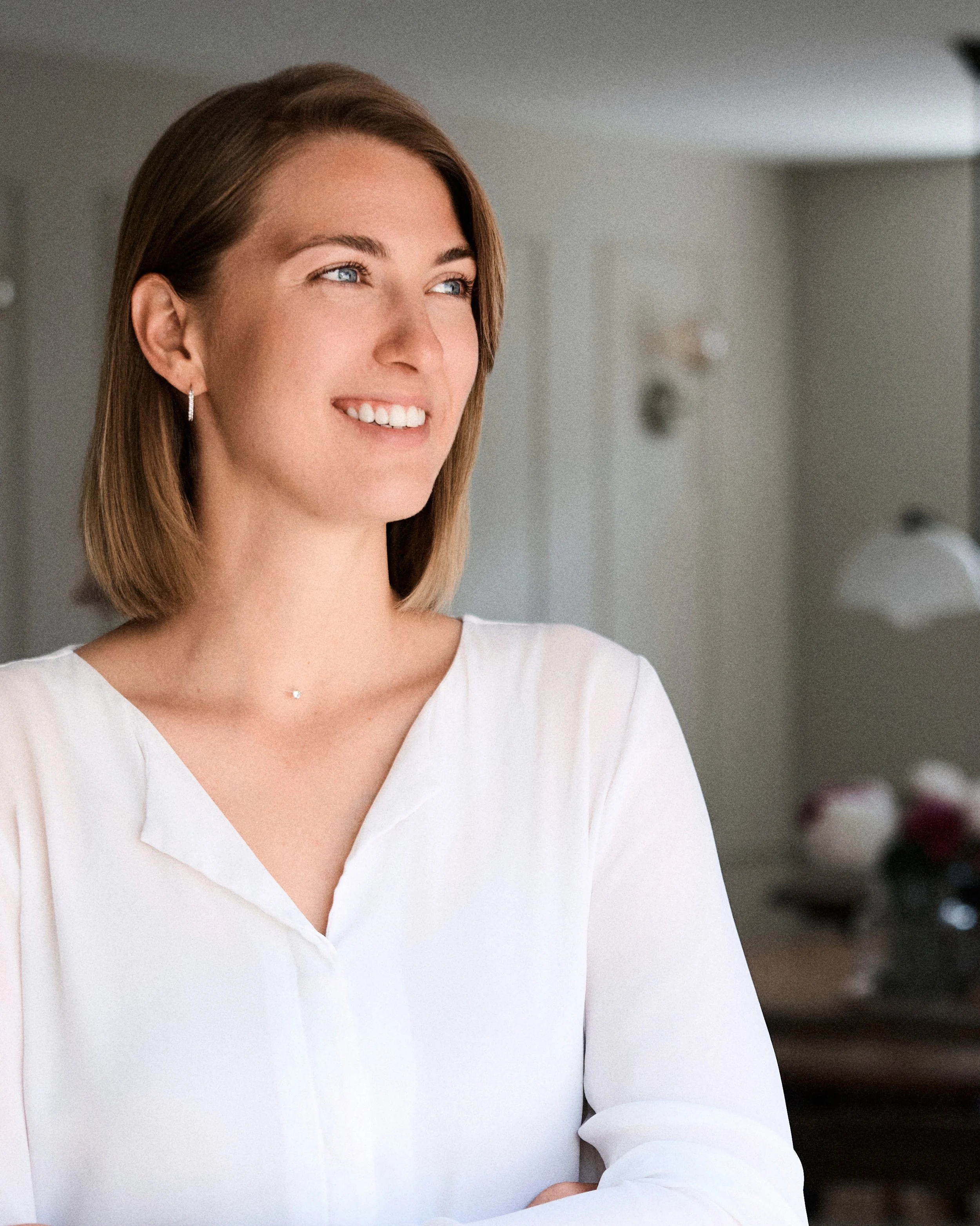 Junge Frau (Vanessa Schaal) mit braunen Haaren und blauen Augen, trägt weiße Bluse, lächelt nachdenklich, im Hintergrund ein Raum mit dunklen Möbeln