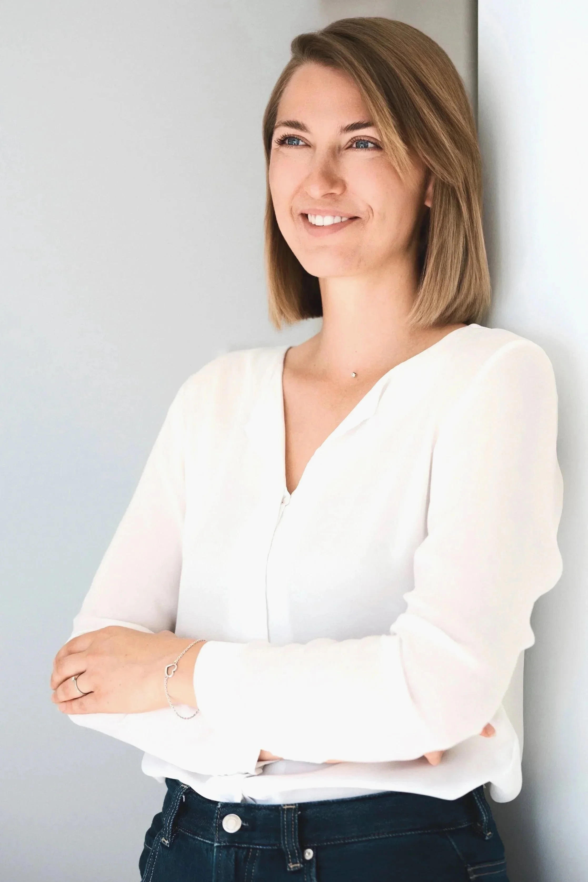 Junge Frau mit braunem, kinnlangem Haar, trägt weiße Bluse und dunkle Jeans, steht lachend an weißer Wand.