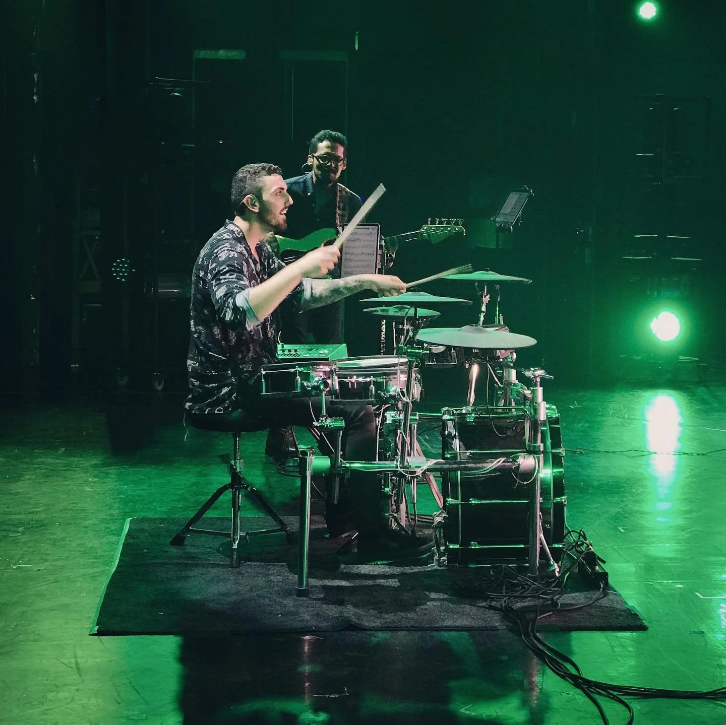 Two musicians performing on stage with green lighting, one sitting at a drum set and the other standing with a guitar.