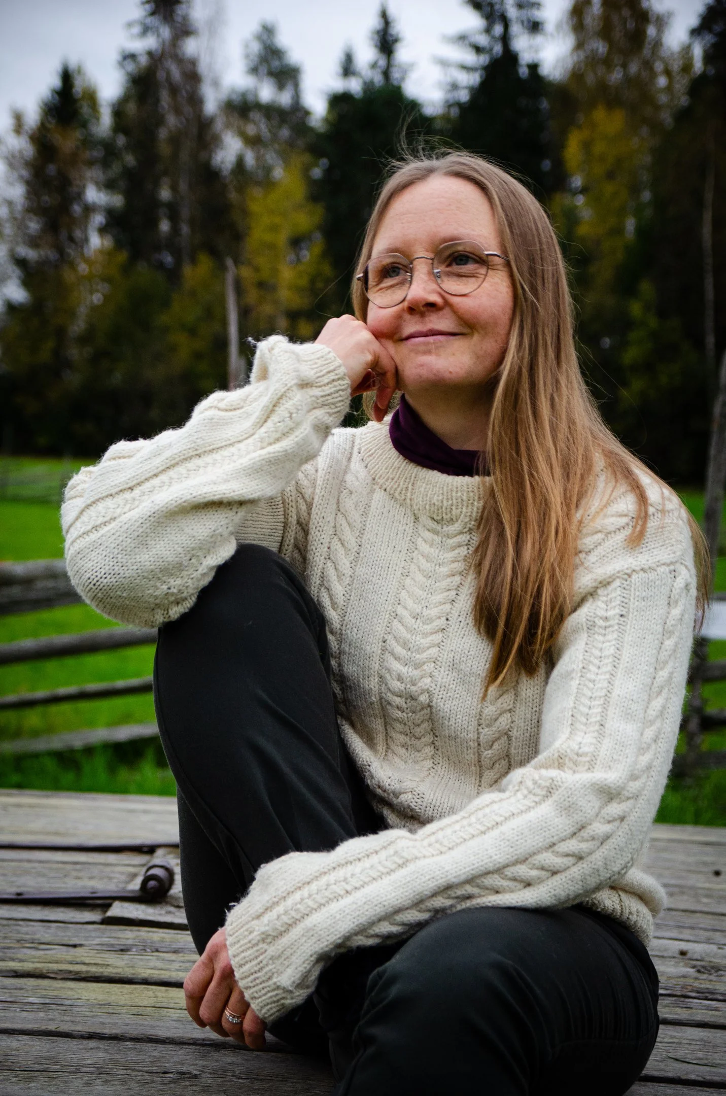 Nainen istuu puistossa, katseli miettimään ja hymyilee, taustalla puita ja vihreää kasvillisuutta.