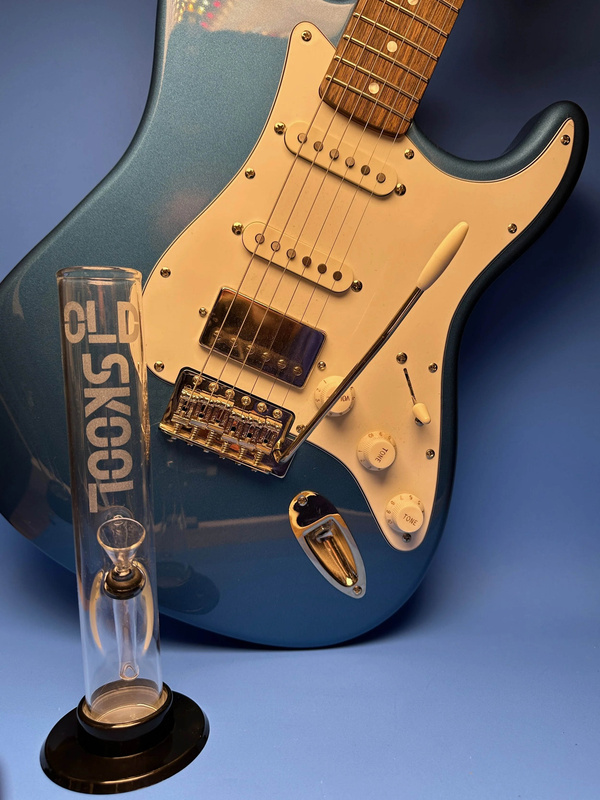 A blue electric guitar with a white pickguard, positioned against a blue background, showcasing a clear glass straight tube water pipe with the "Old Skool" logo etched on the front of it.