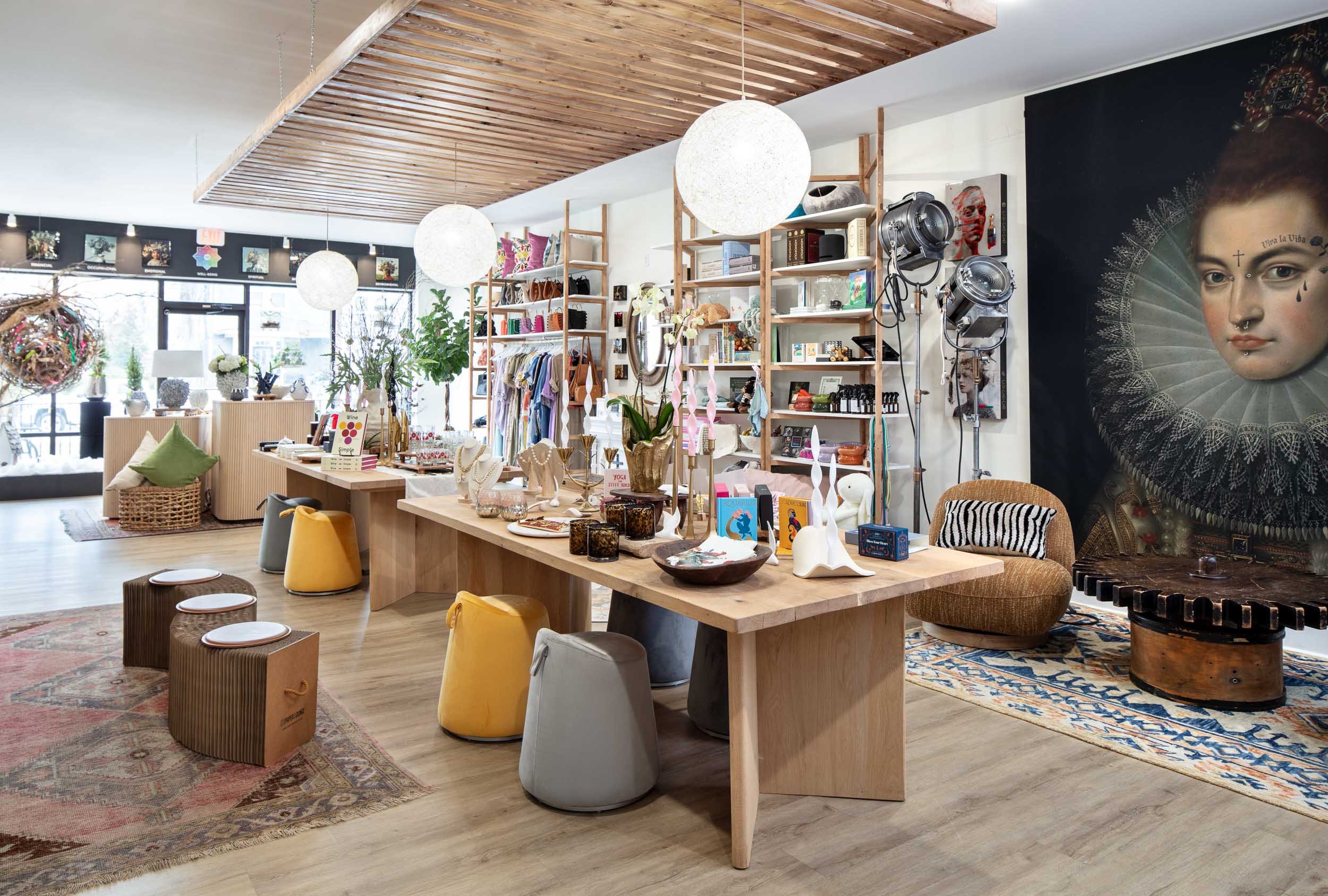 Interior of a boutique shop featuring wooden display tables with decorative items, clothing racks, and a seating area with stools and a cushioned chair, decorated with plants, artwork, and hanging globe lights.