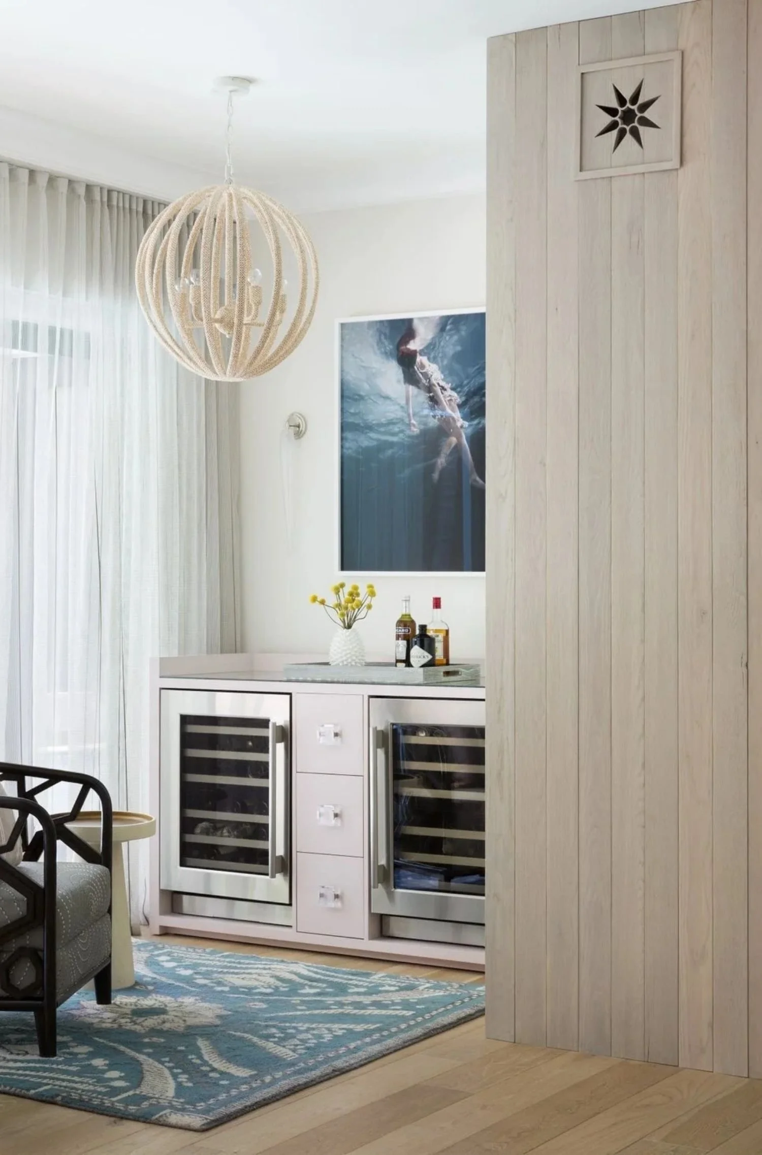 A sitting room with a wine cooling fridge. A piece of art showing someone swimming in dark blue waters sits above the small bar. A blue rug creates additional contrast against the light neutral shades of the wood panelling and sheer draperies.