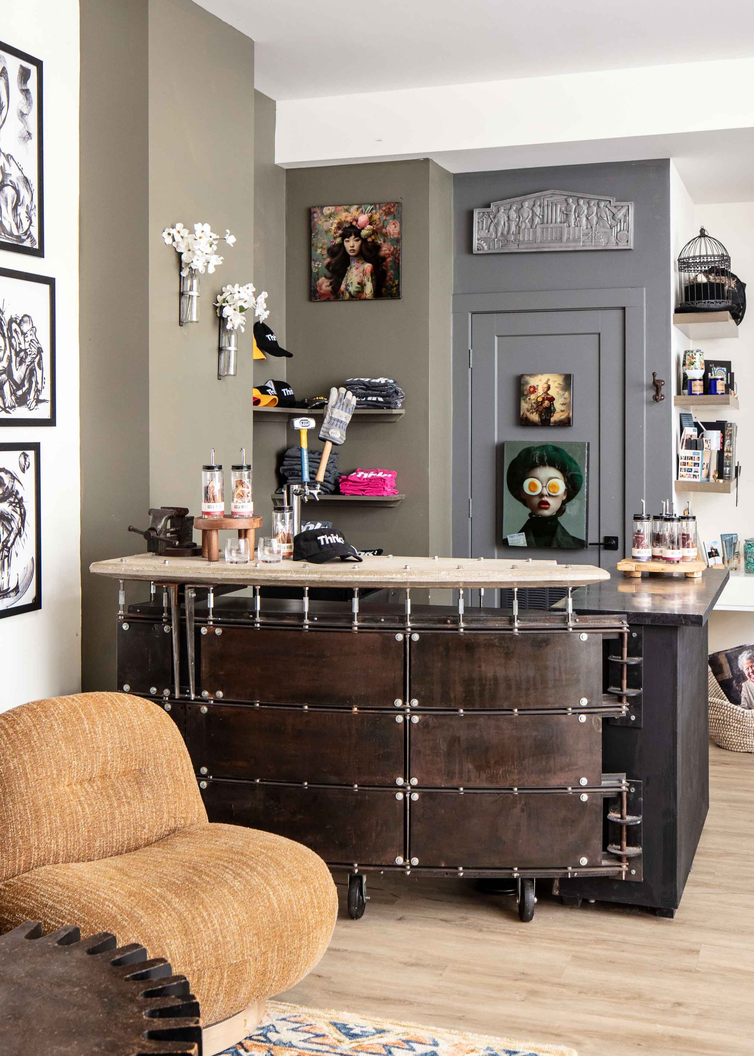 Interior of a cafe or boutique with a rustic dark wood counter, decorated with glasses and small plants, shelves with hats, folded clothes, artwork on the walls, a tan upholstered chair, and various decorative items.