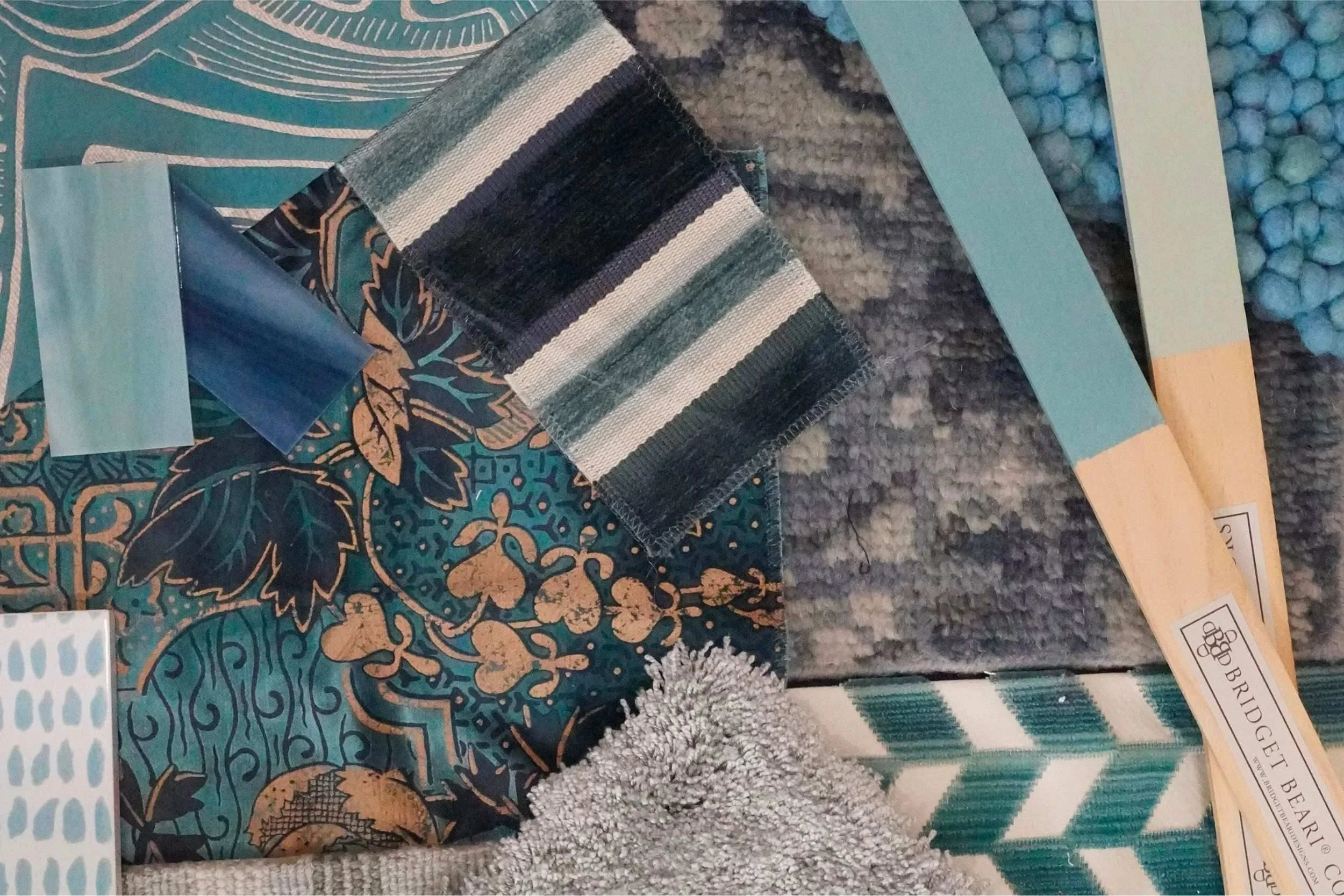An array of patterned blue fabrics and rug samples laid out. 2 light blue paint color sticks are laid on the right.