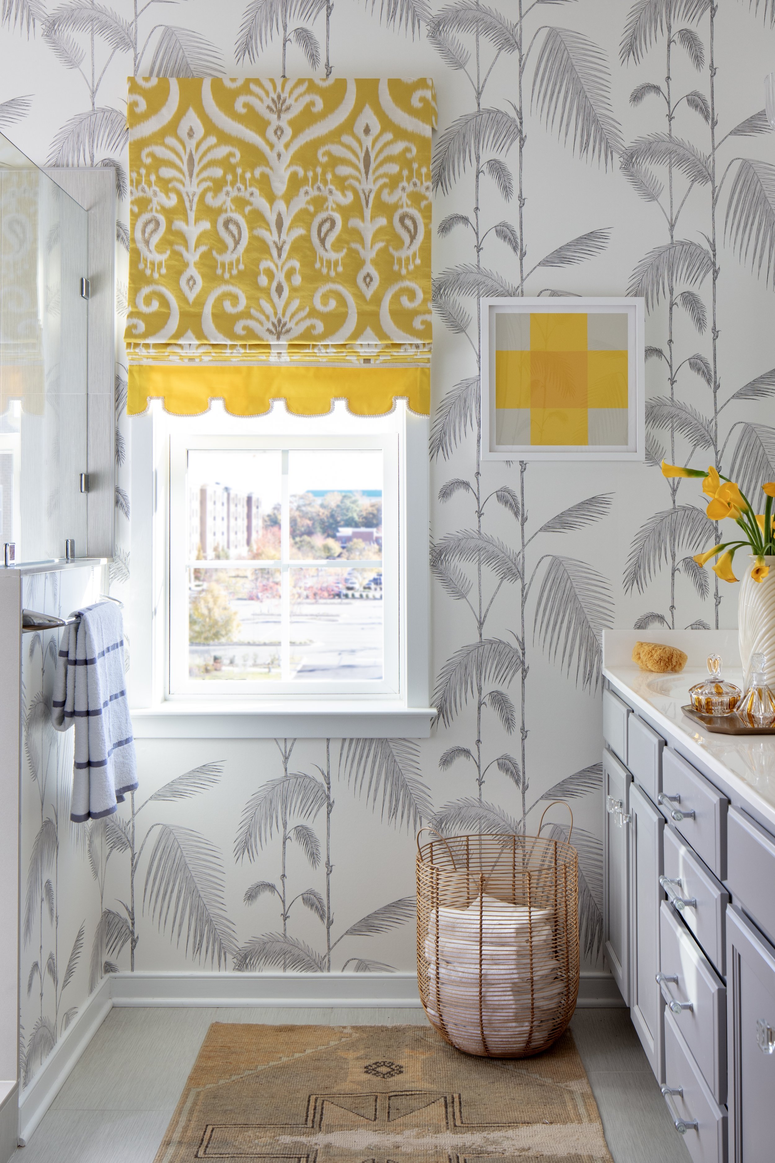 Bright bathroom with gray palm tree wallpaper, yellow and white window shade, white cabinetry, and a woven laundry basket.