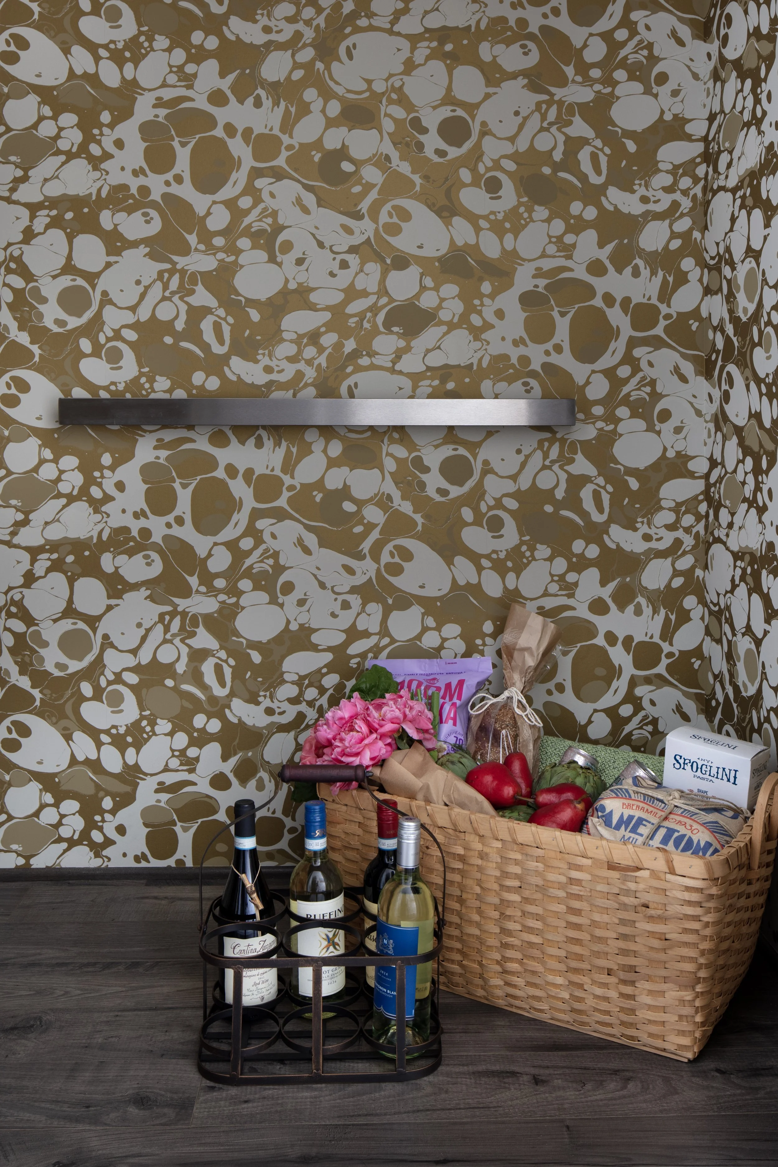 A basket of groceries containing flowers, apples, and packaged goods, next to a wine rack with three bottles, in a room with a patterned gold and white wall and dark wooden floor.