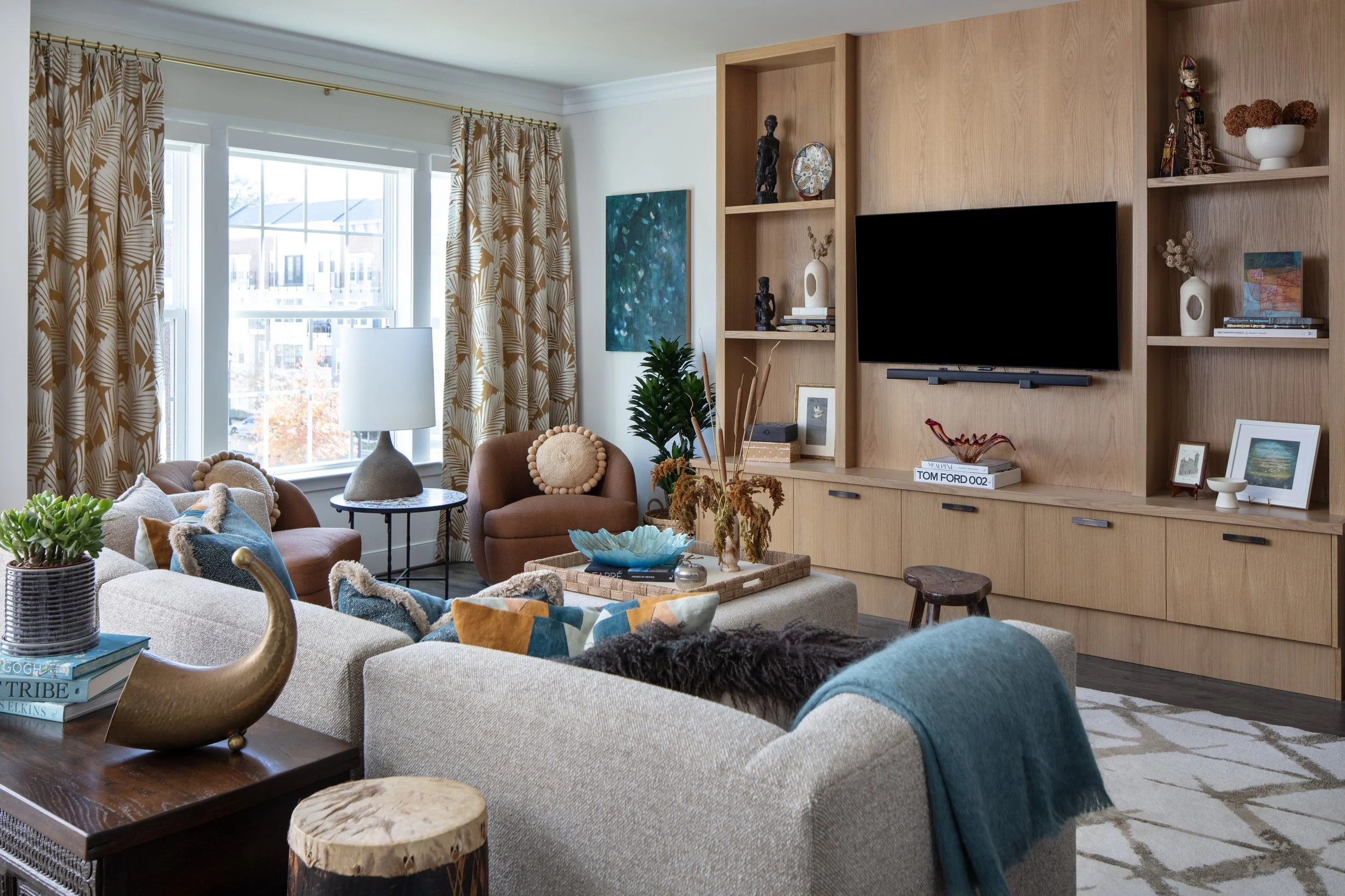 Living room with large windows, beige sofa with colorful pillows, two brown armchairs, wooden entertainment center with TV, and various decorative items and plants