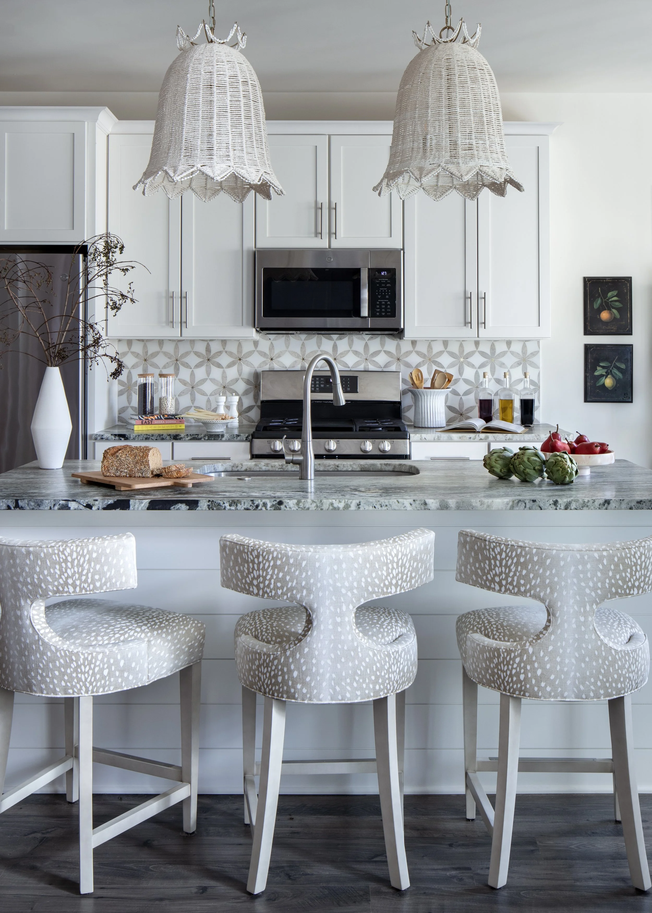 Modern kitchen with white cabinets, pendant lighting, microwave oven, stovetop, decorative backsplash, and three patterned barstools at a marble counter.