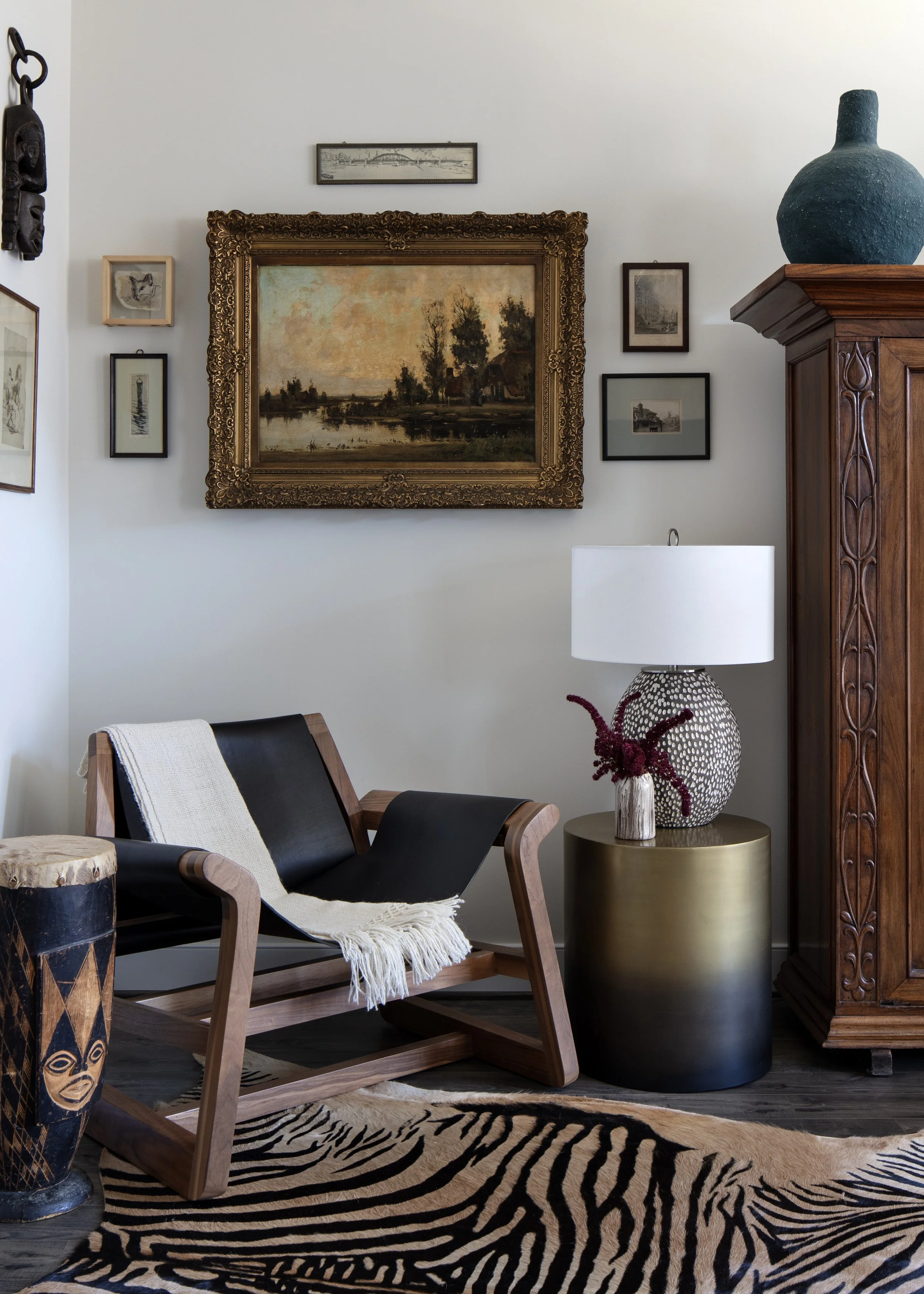 Living room corner with a black leather and wood lounge chair, zebra-patterned rug, a gold side table with a white lamp and floral decor, a wooden wardrobe, and framed artwork on the white wall.