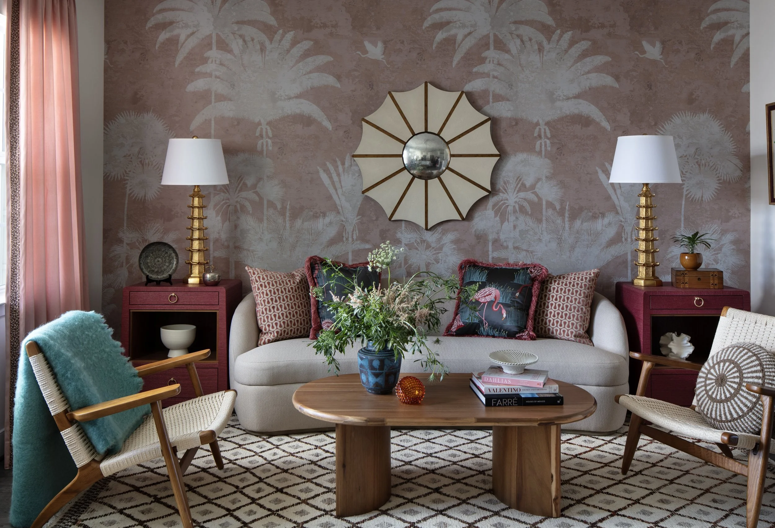 Living room with a beige couch, mid-century modern chairs, and pink side tables with lamps. Decor includes a wall mirror, a floral arrangement, pillows, and books.
