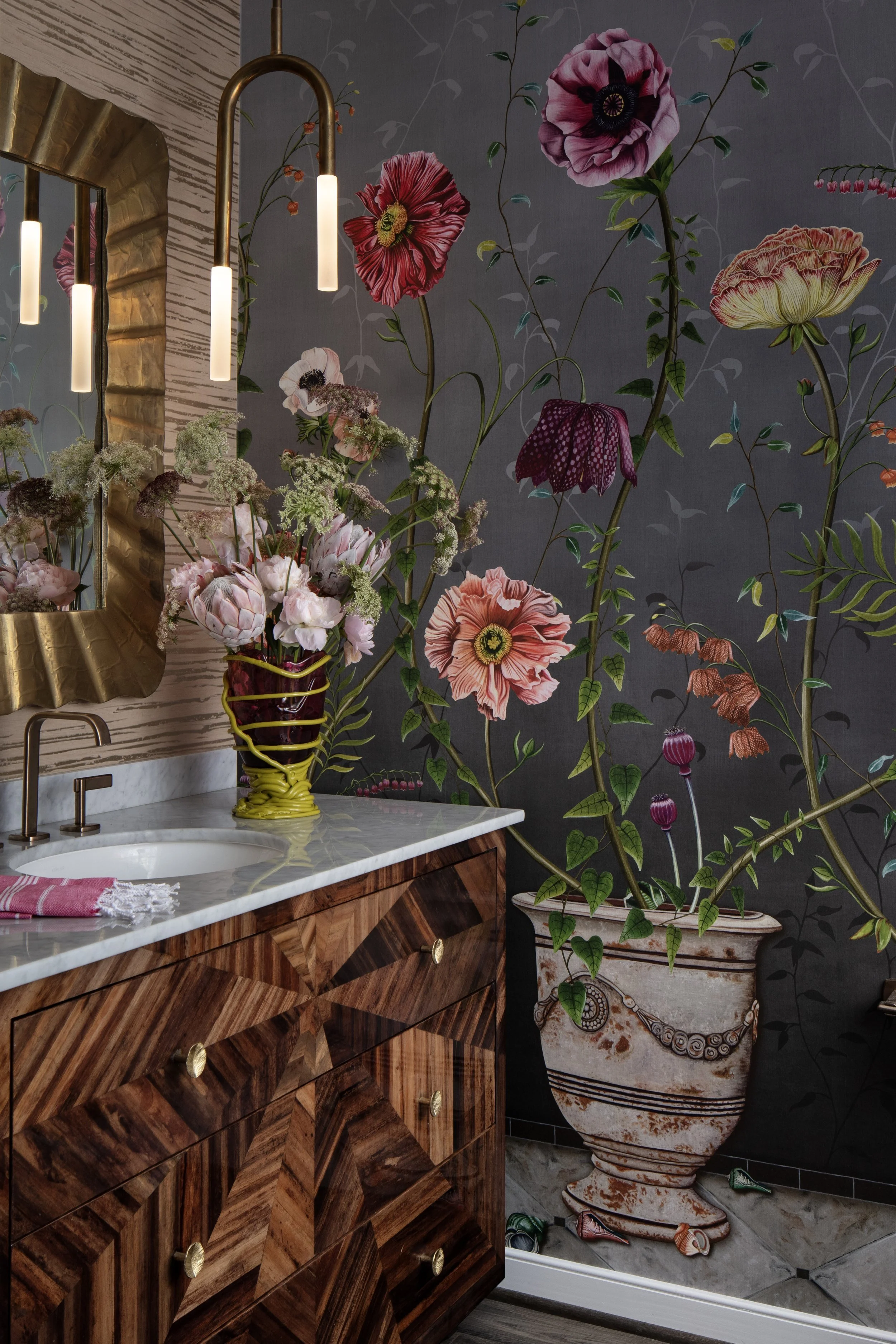 Bathroom with floral wallpaper, a mirror, a marble countertop with a vase of flowers, and a large decorative flowerpot with blooming plants.