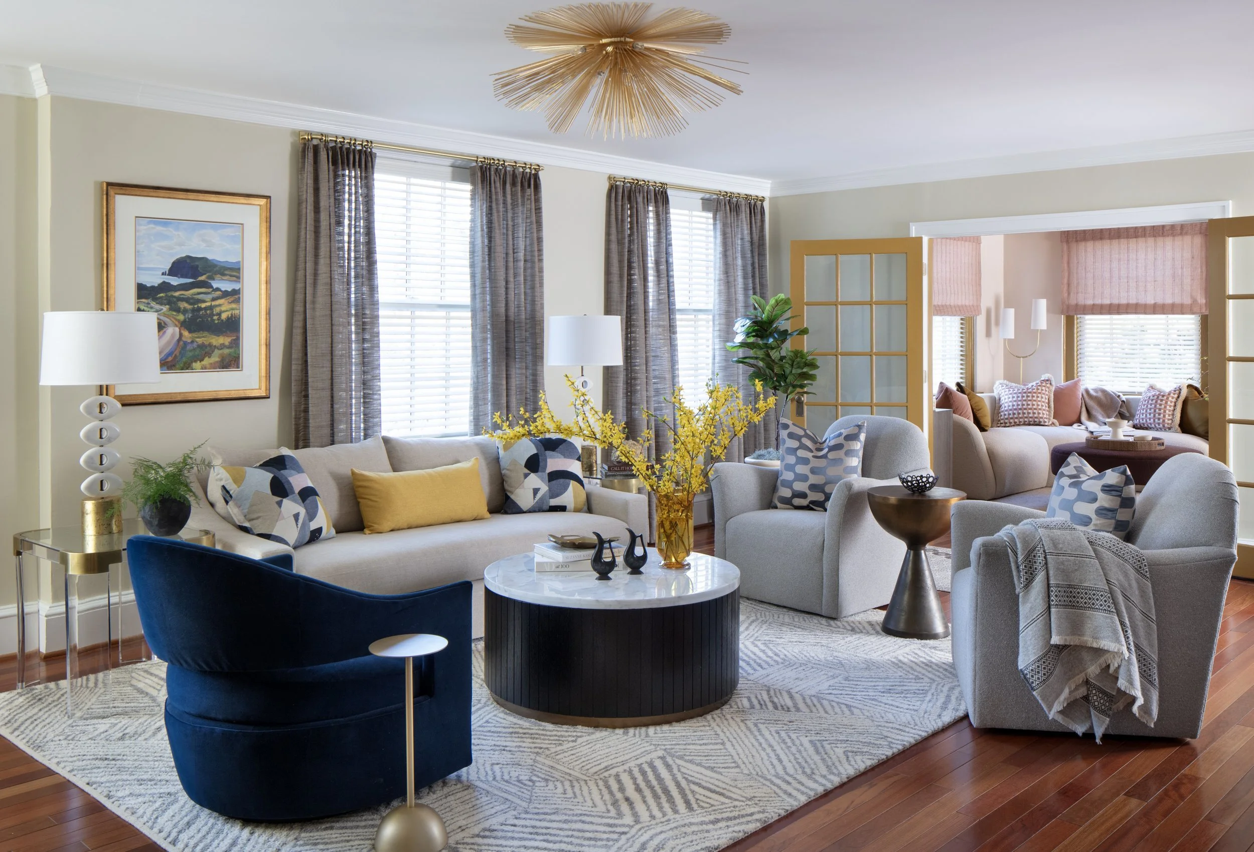 Elegant living room with beige sofas, patterned cushions, a blue velvet armchair, and a black round coffee table with yellow flowers in a glass vase. Large windows with gray curtains, wooden flooring, and a decorative ceiling light. A glass-paneled room divider opens into a sunroom with pink shades and additional seating.
