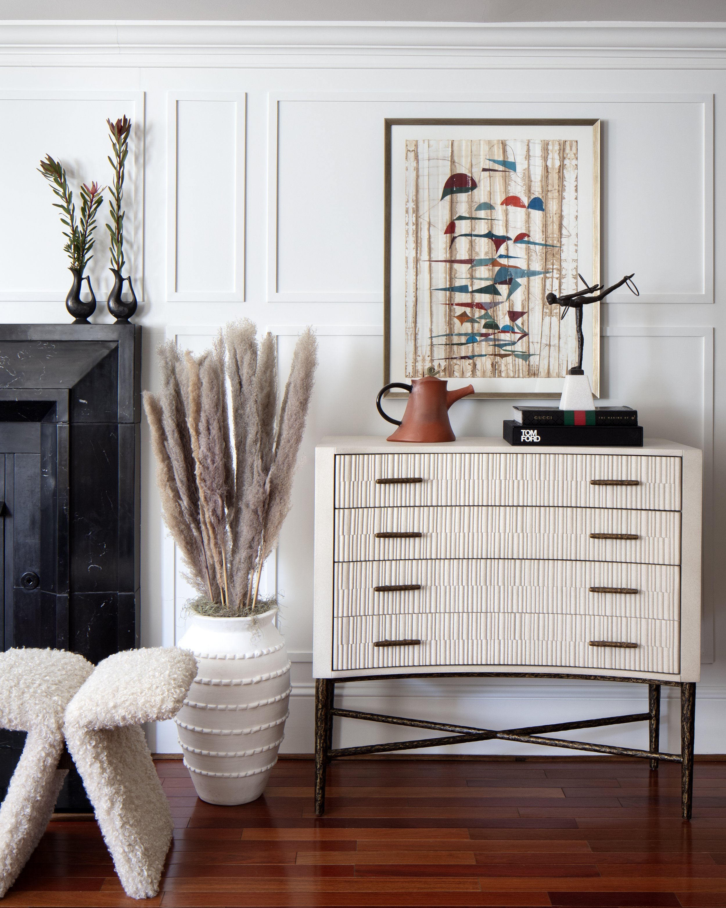 Interior living room decor with a black fireplace, white wainscoting walls, a white textured sideboard, framed abstract art, a terracotta watering can, a sculpture, and a large vase with pampas grass.