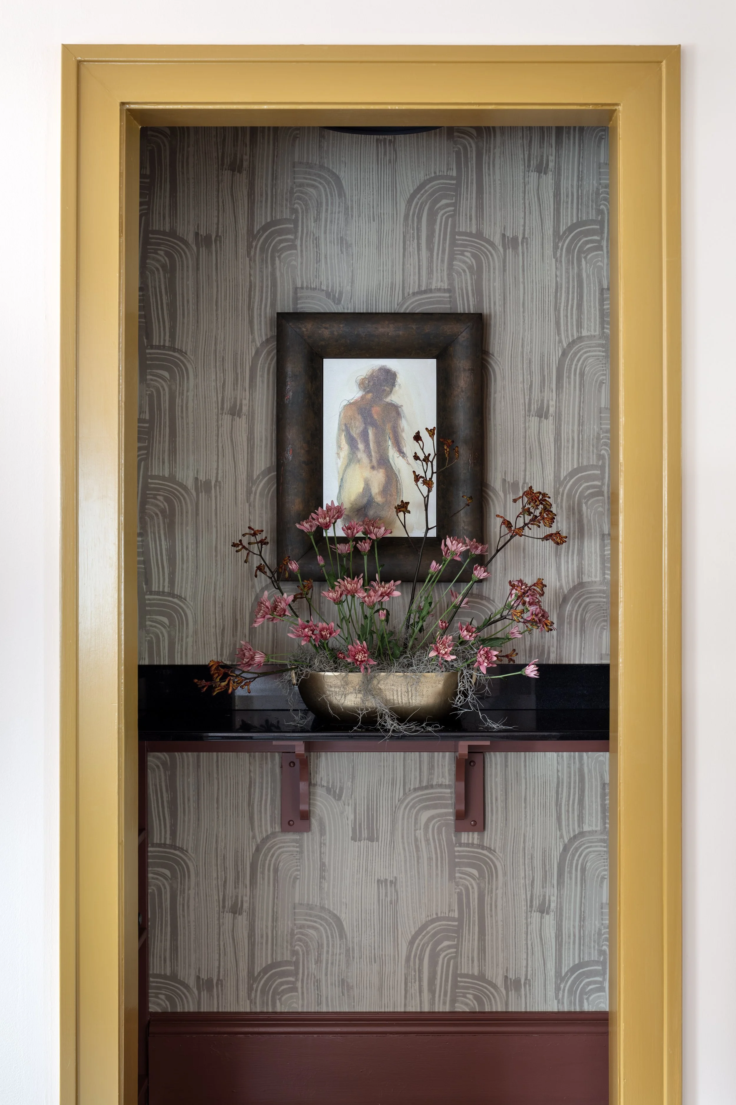 A decorative niche with a gold frame, featuring a framed painting of a nude woman, a black shelf with pink flowers and moss, and patterned wallpaper in the background.