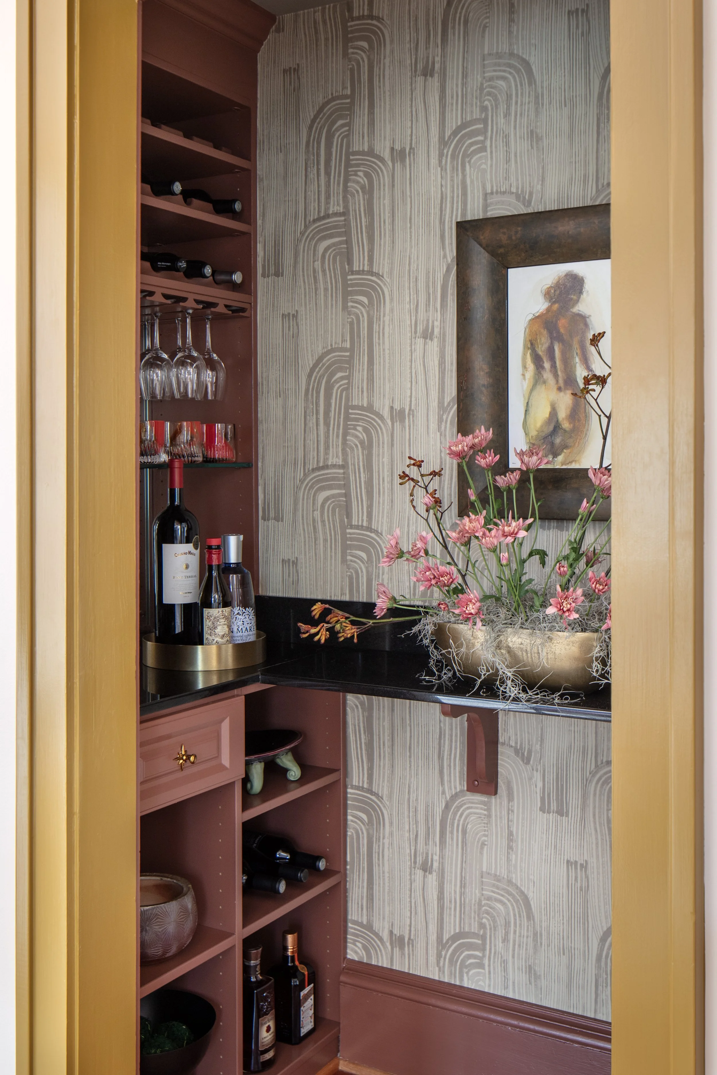 A small interior space with pink built-in shelves holding wine bottles, glasses, and bottles of liquor. There is a dark countertop with a large floral arrangement in a golden bowl, pink flowers with green stems, and a framed artwork of a nude woman o