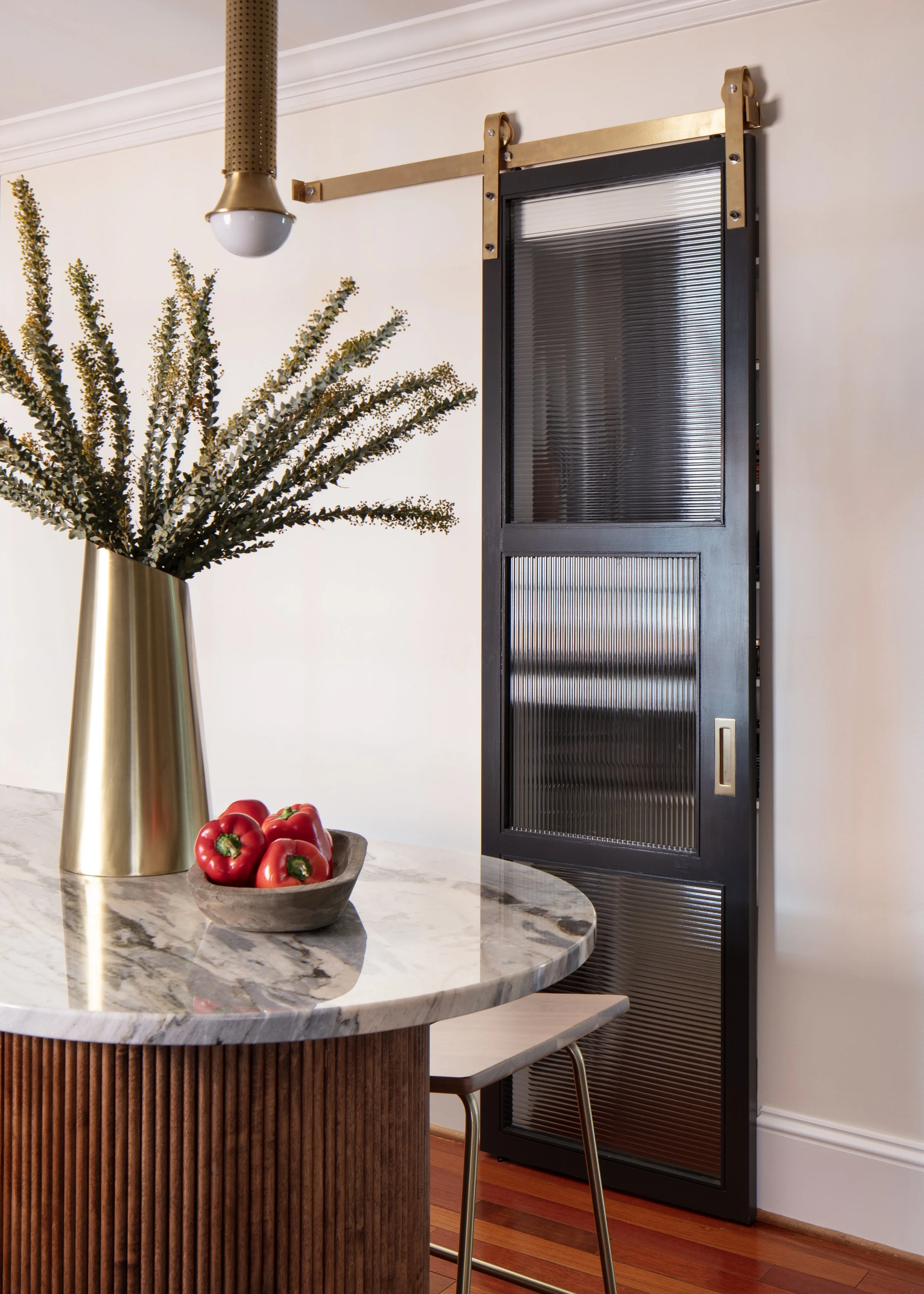 Modern interior with a marble-topped table, a gold vase with green foliage, a bowl of red peppers, and a sliding black louvered door.