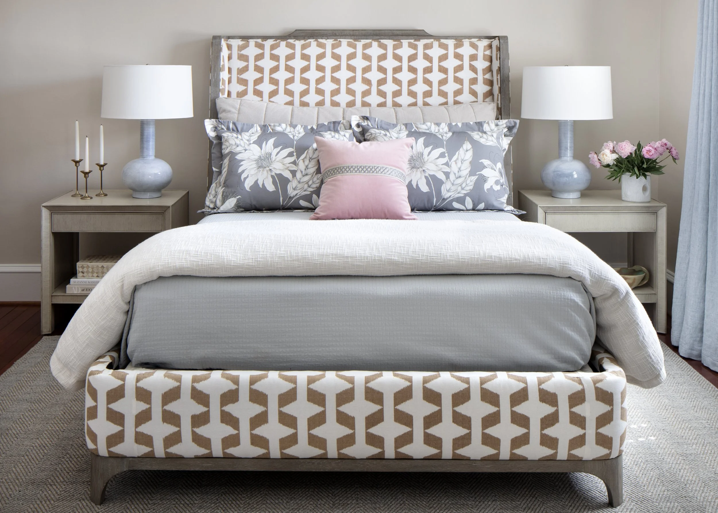 A bedroom with a bed, nightstands, lamps, and decorative pillows. The bed has a geometric patterned headboard and footboard, with floral pillows and a pink accent pillow. The nightstands hold lamps, with one side featuring a vase of pink flowers.