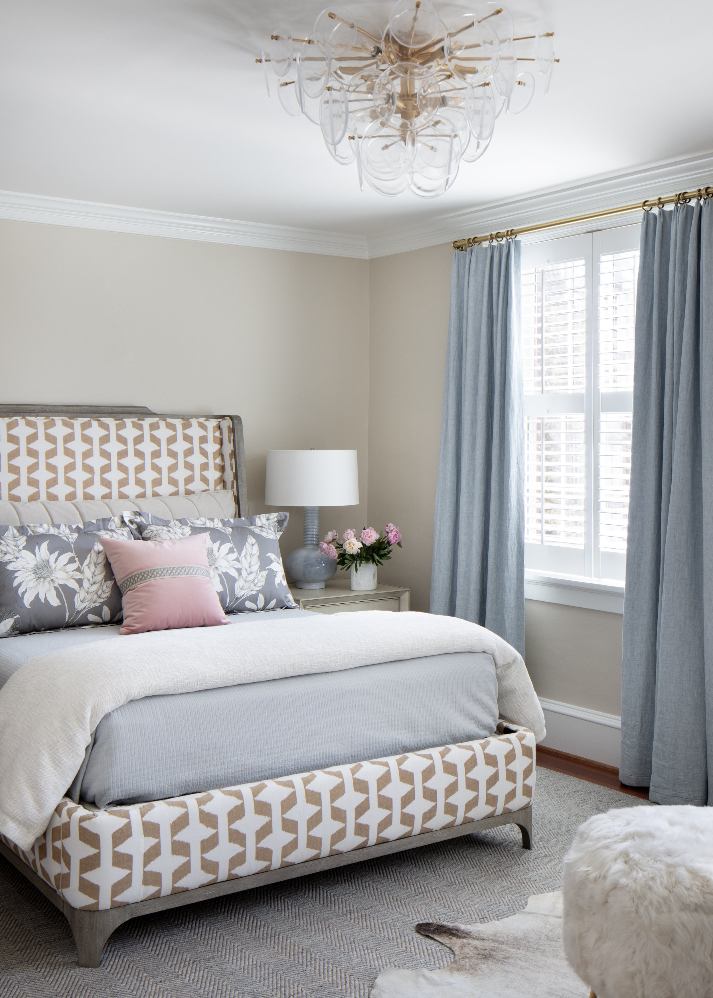 A bedroom with a bed that has a patterned headboard, gray and pink pillows, and a cream throw blanket. A nightstand with a white lamp and a vase of pink flowers is next to the bed. Large window with blinds and blue curtains. A modern chandelier hangs