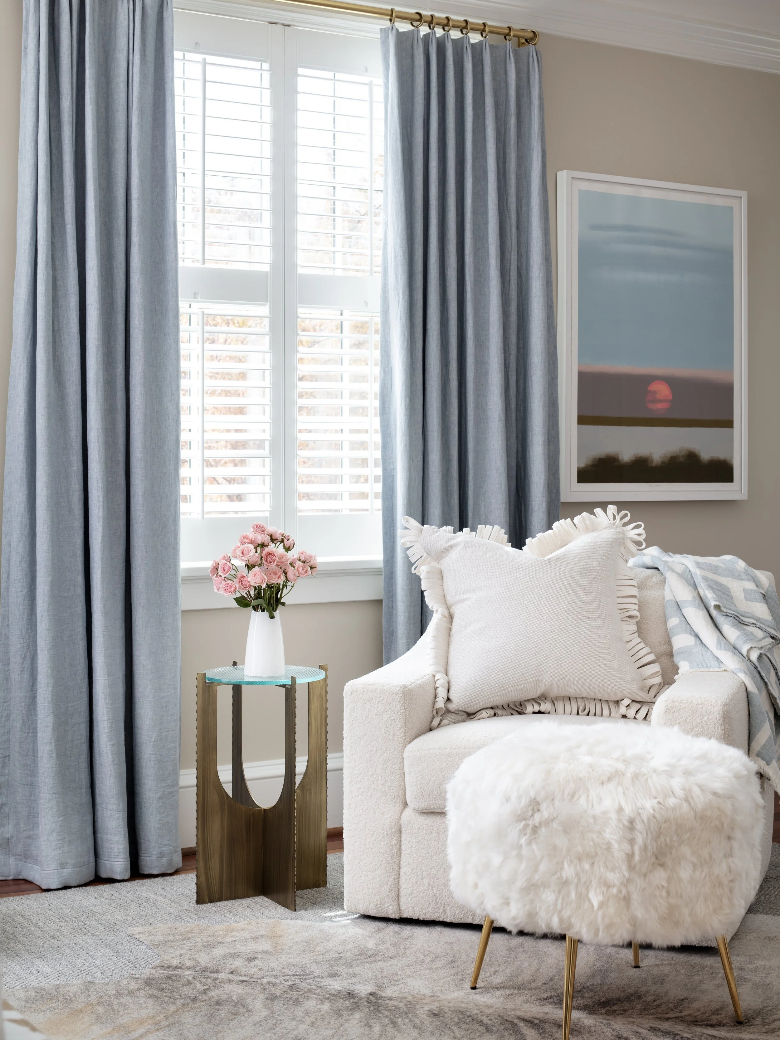 Living room with light curtains, a white couch with pillows, a fluffy white ottoman, a side table with pink roses, and a framed art print of a sunset.