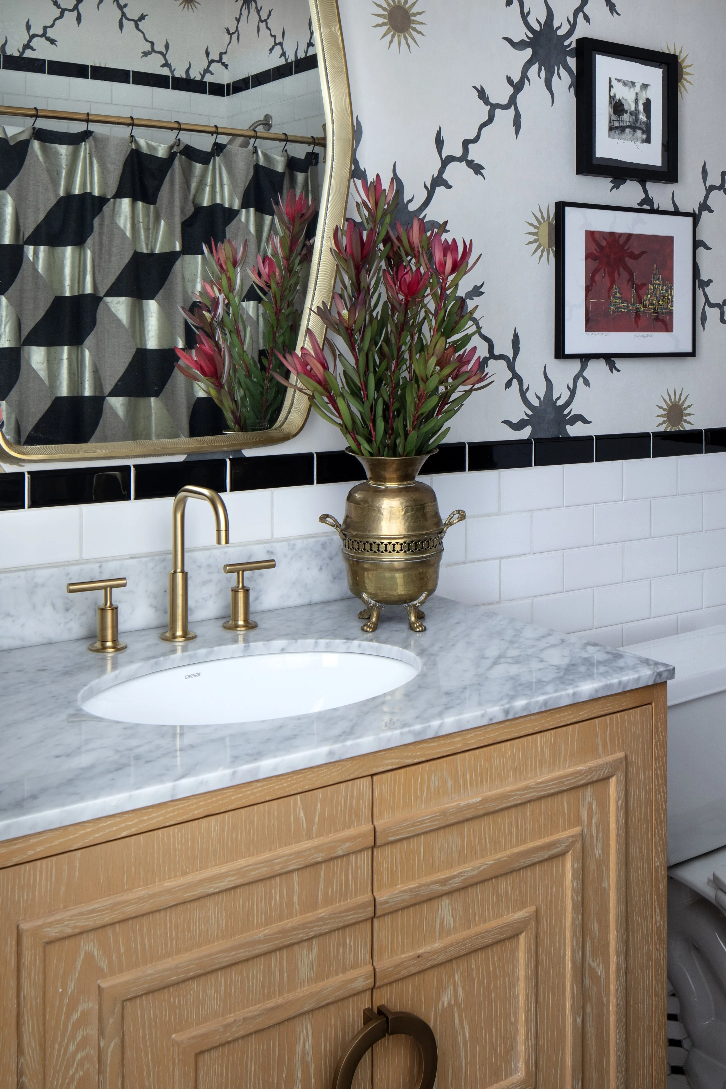 Bathroom sink with gold fixtures and a marble countertop, a tall brass vase with red flowers, a mirror with a gold frame, framed artwork on the wall, a shower curtain with a black and beige geometric pattern, and a section of a toilet.