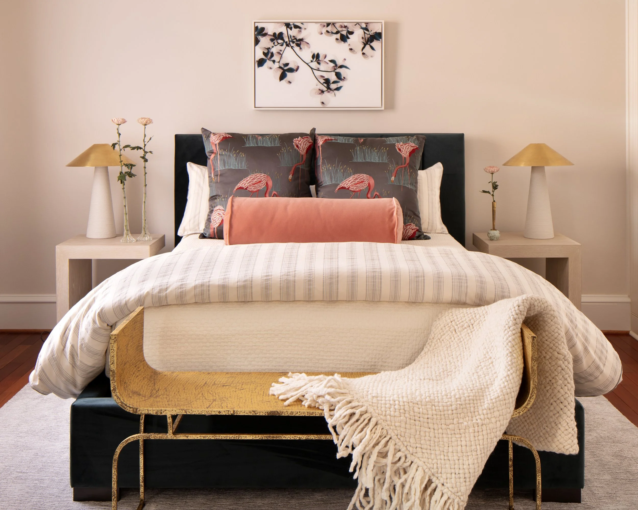 A neatly made bed with flamingo-patterned pillows, a coral-colored bolster, with two nightstands with lamps and decorative items, and a framed botanical artwork above the headboard in a bedroom.