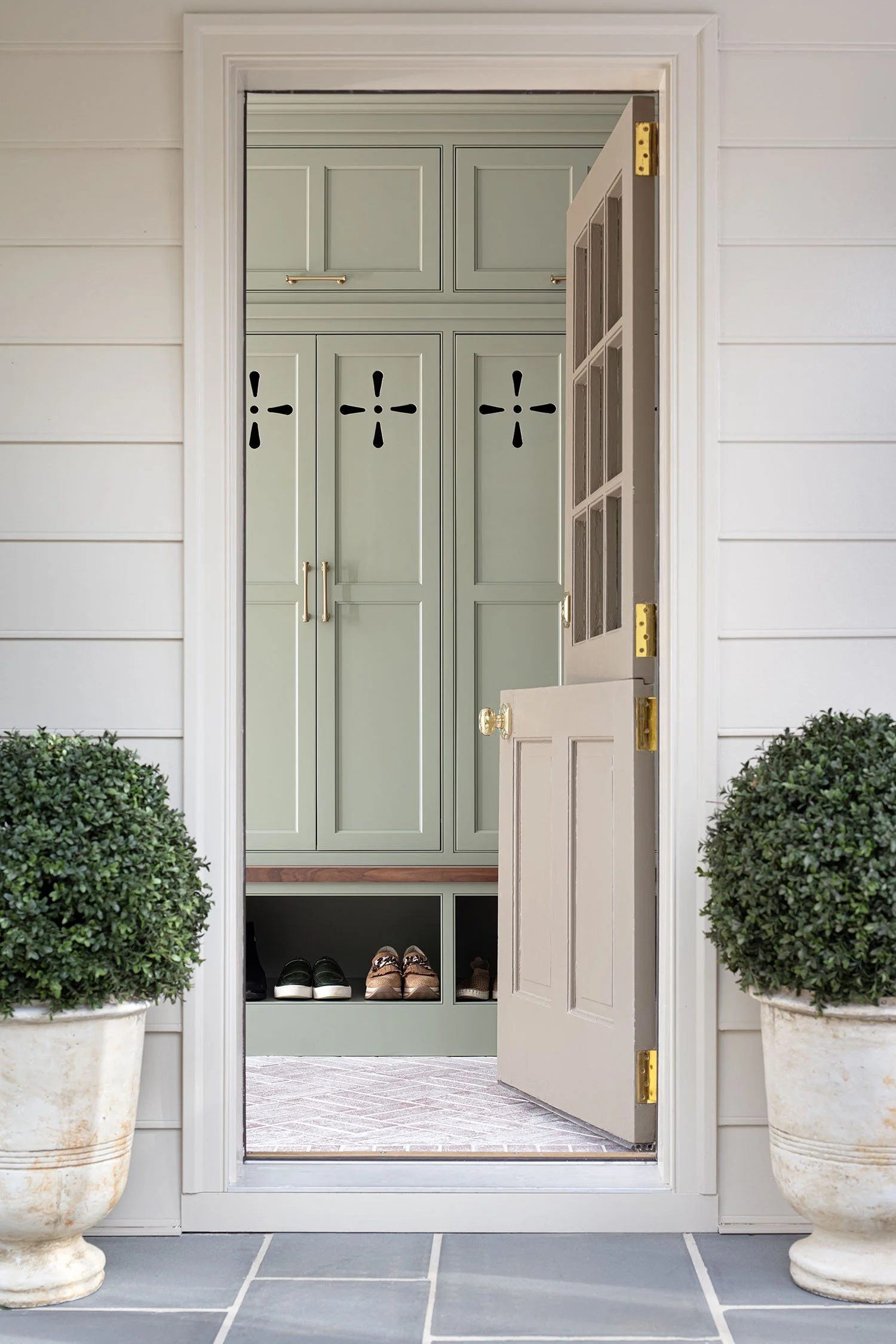 Open front door of a house, revealing a green closet with storage cabinets and shoes inside, flanked by two potted shrubs outside.