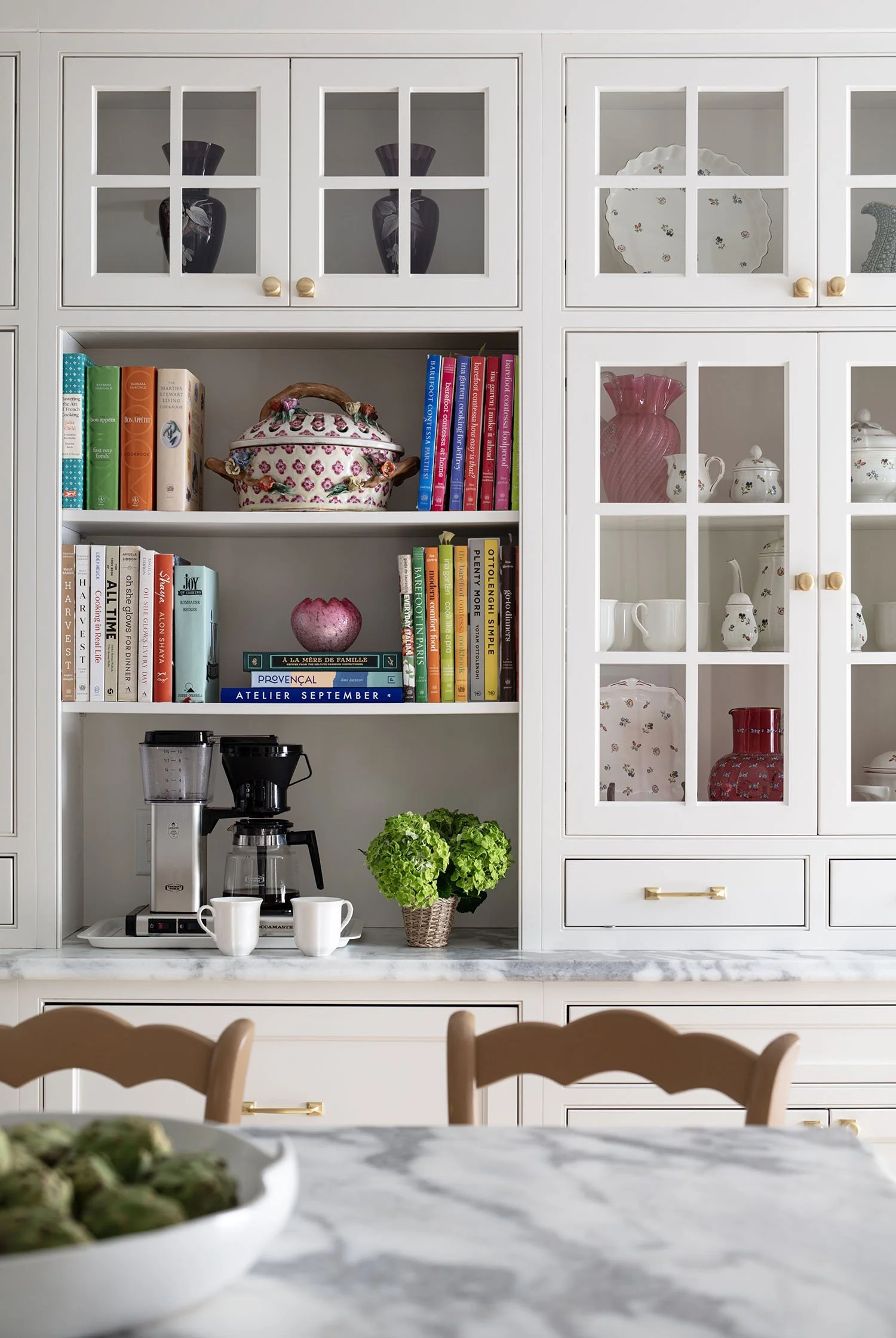 Kitchen cabinet and countertop with coffee maker, two coffee mugs, green plant in basket, and a marble table with a bowl of artichokes.