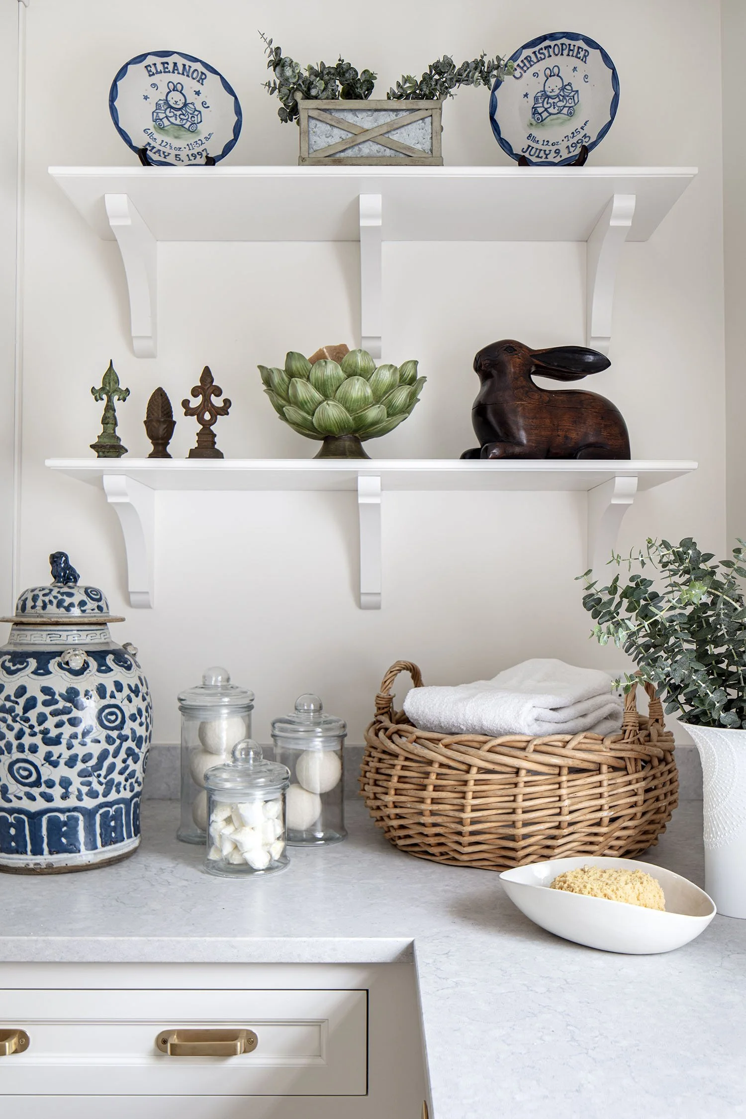 Decorative kitchen shelf with Easter-themed pottery, a wooden bunny sculpture, green artichoke centerpiece, small statues, glass jars with candies, a wicker basket with folded towels, a large blue and white jar, a white bowl with a sponge, and a vase