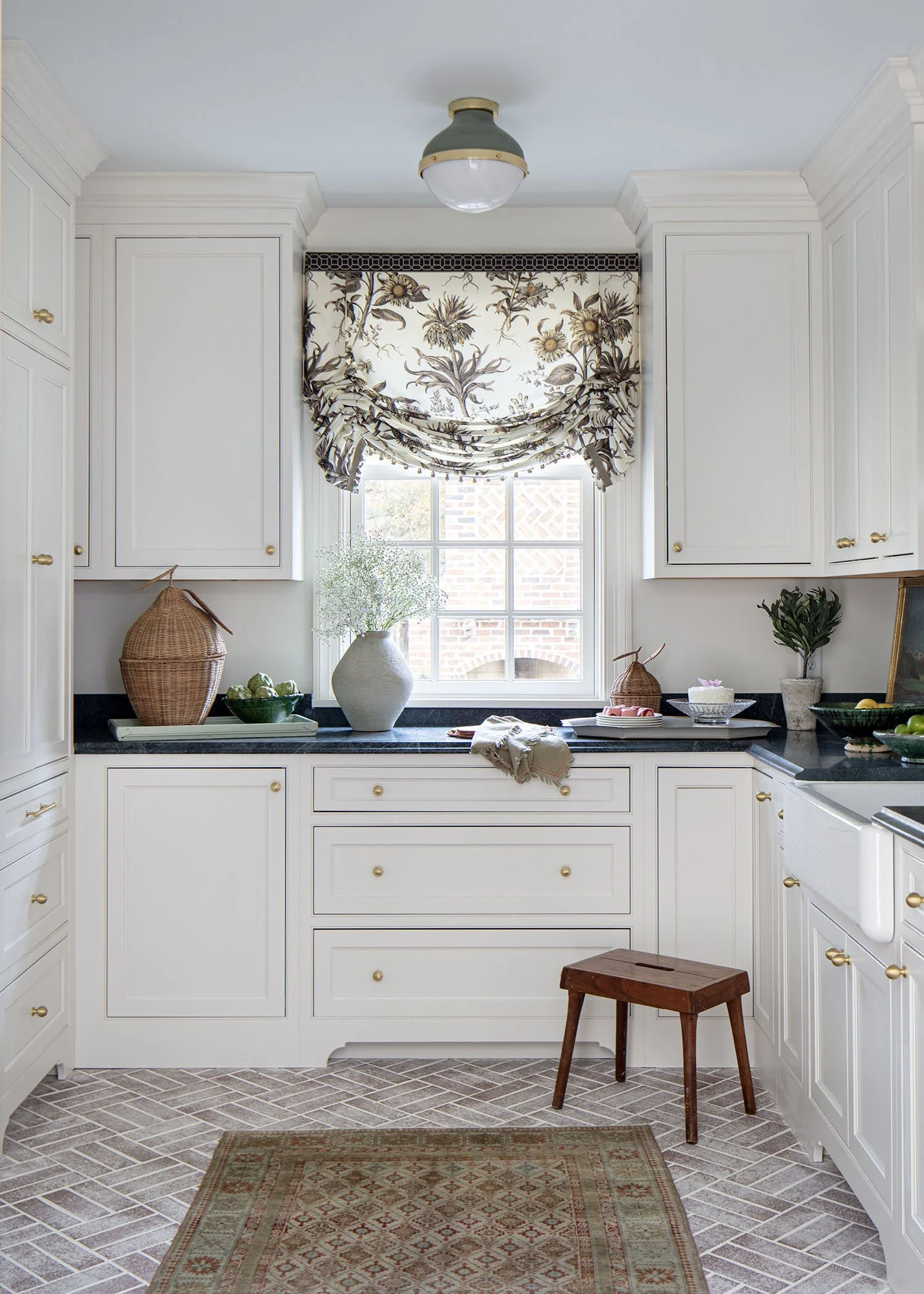 Bright kitchen with white cabinets, black countertops, and a small wooden stool. Decorative items include a potted plant, a wicker jar, a large ceramic vase with baby's breath flowers, and a tray of apples. A window with floral-patterned curtains all