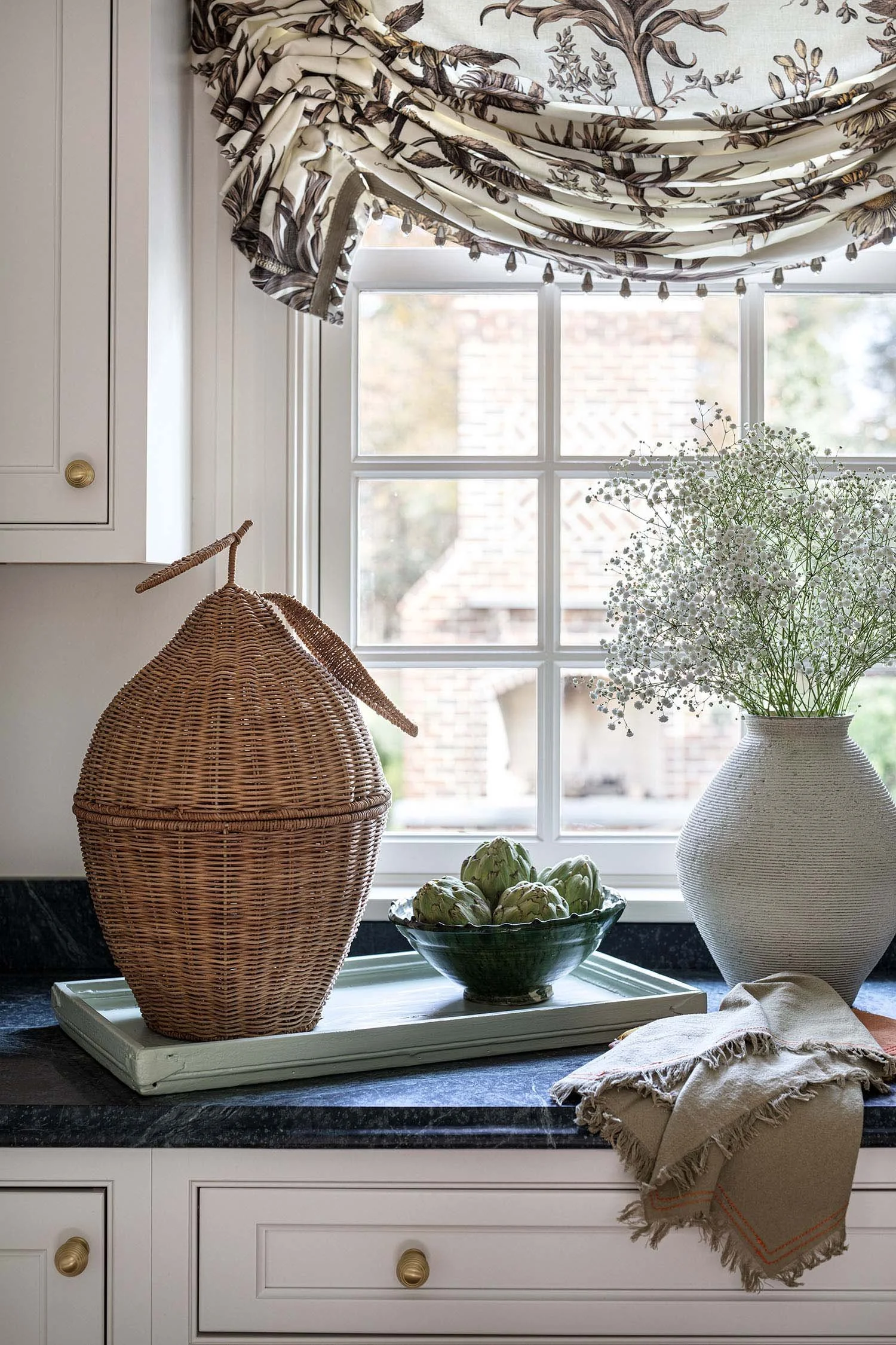Decorative kitchen window sill with a wicker container, a bowl of artichokes, a large white vase with baby's breath flowers, part of a cream-colored cabinet with gold knobs, and a beige cloth napkin.
