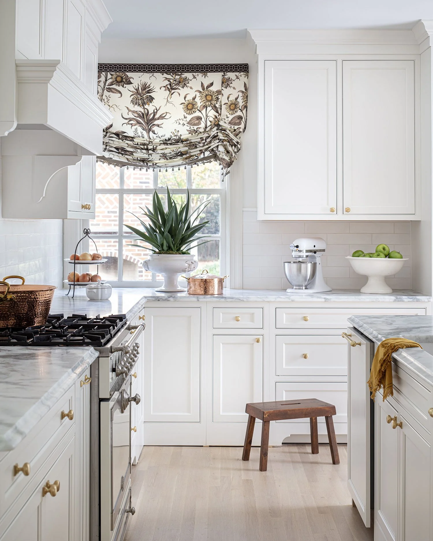 Bright white kitchen with marble countertops, a window with a floral Roman shade, a large potted plant, a stand holding apples, a bowl of green apples, a stand mixer, a copper pot, a small wooden stool, and cream-colored cabinets with gold knobs.