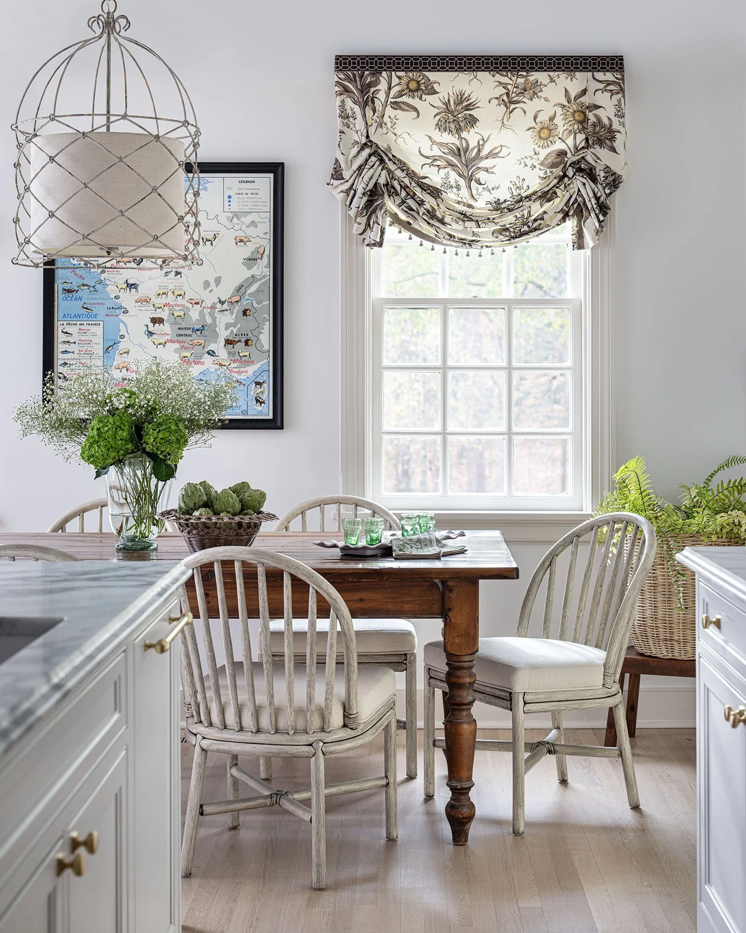 A bright dining area with a wooden table, four white wooden chairs, a floral window valance, a framed animal map on the wall, a view of the outdoors through a large window, and a bouquet of green flowers on the table.