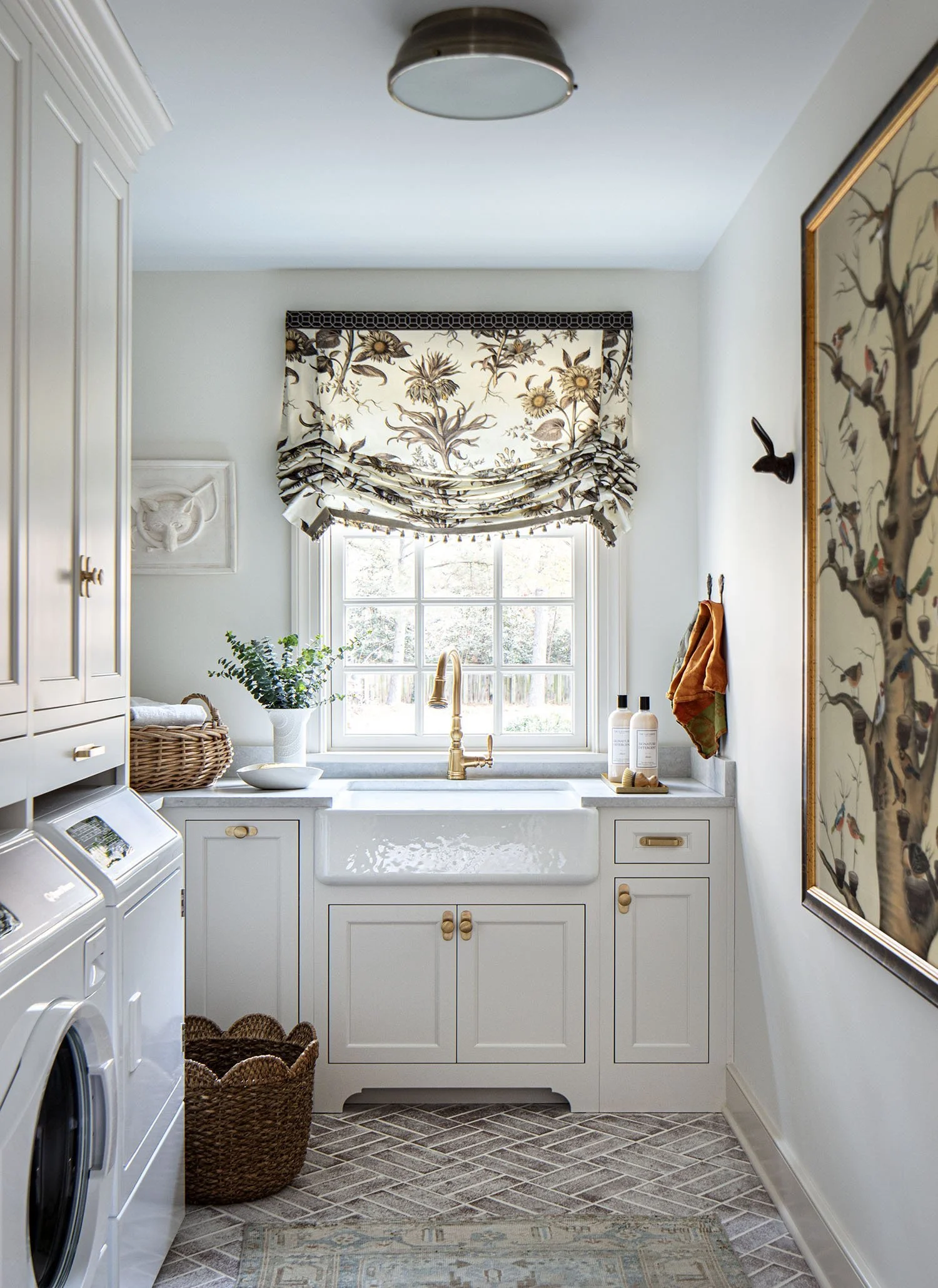 A laundry room with a white utility sink beneath a window with floral roman shades, cabinets, a washing machine, wicker basket, and wall decorations.