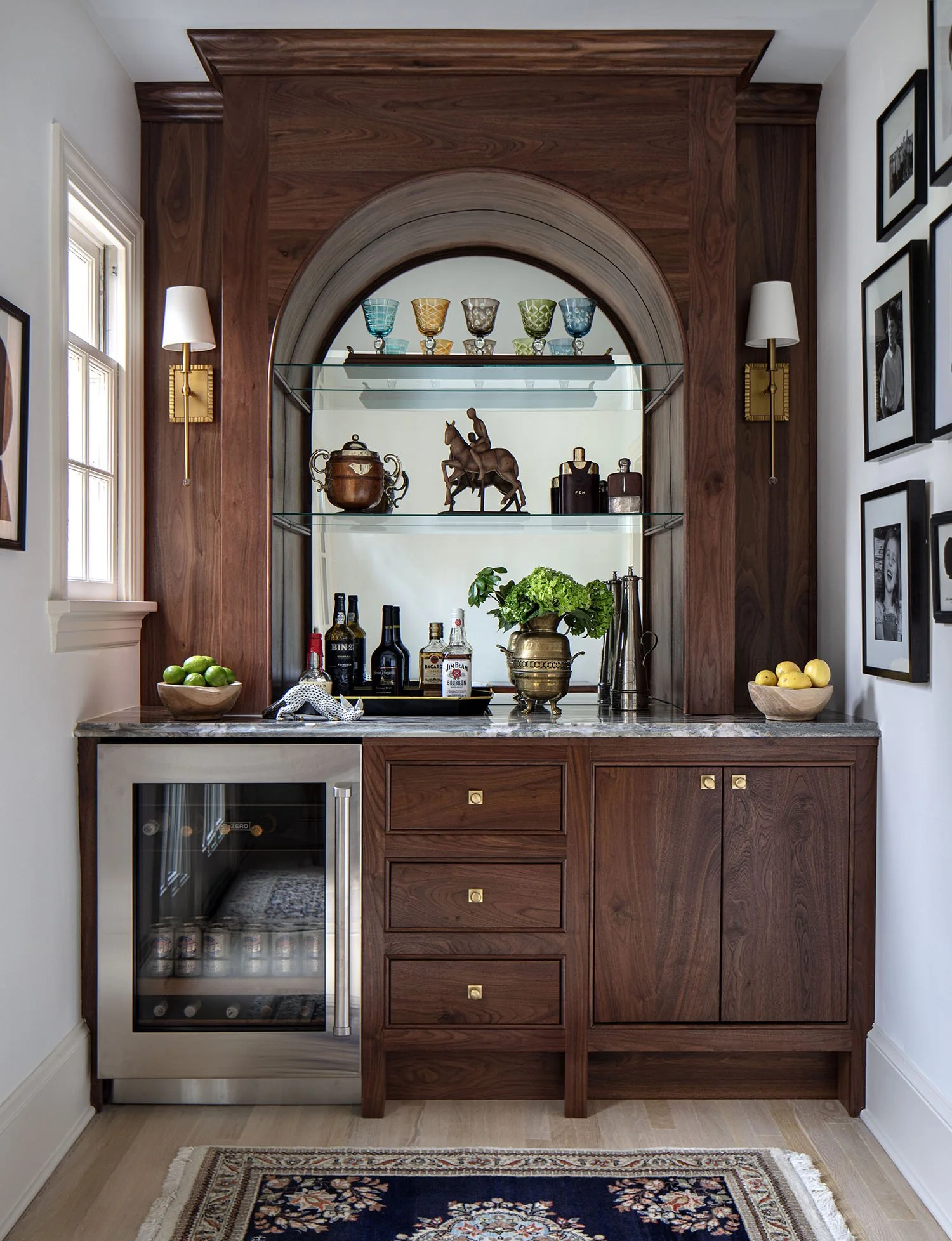 A small wet bar with a wooden cabinet, glass shelving, colorful drinkware, bottles, and decorative items. There's a mini fridge or wine cooler on the lower left and a white window on the left side.