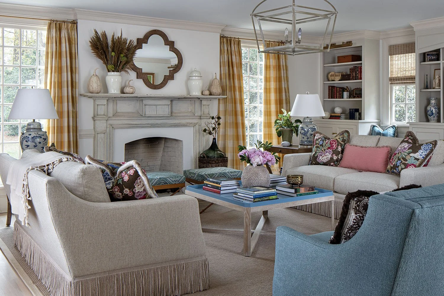 Living room with beige and blue sofas, floral pillows, a blue coffee table, a white fireplace, yellow checkered curtains, and decorative vases and plants.