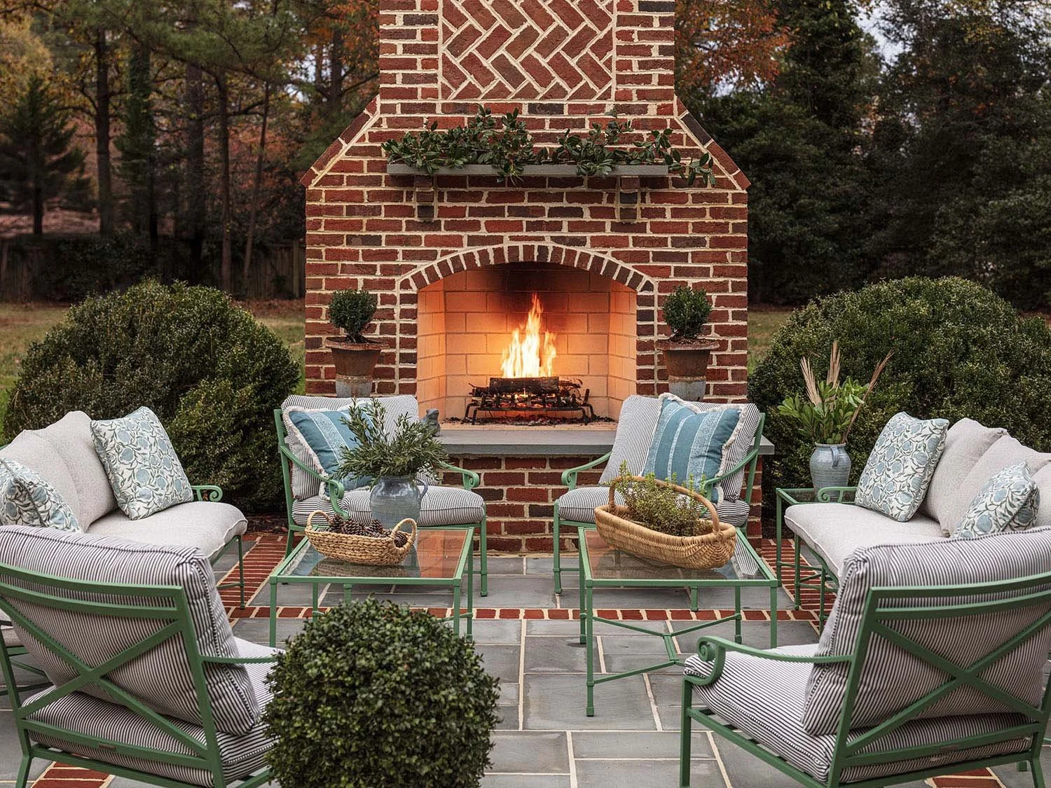 Cozy outdoor patio with green metal furniture and cushions, around a brick fireplace with a fire burning, surrounded by bushes and trees.