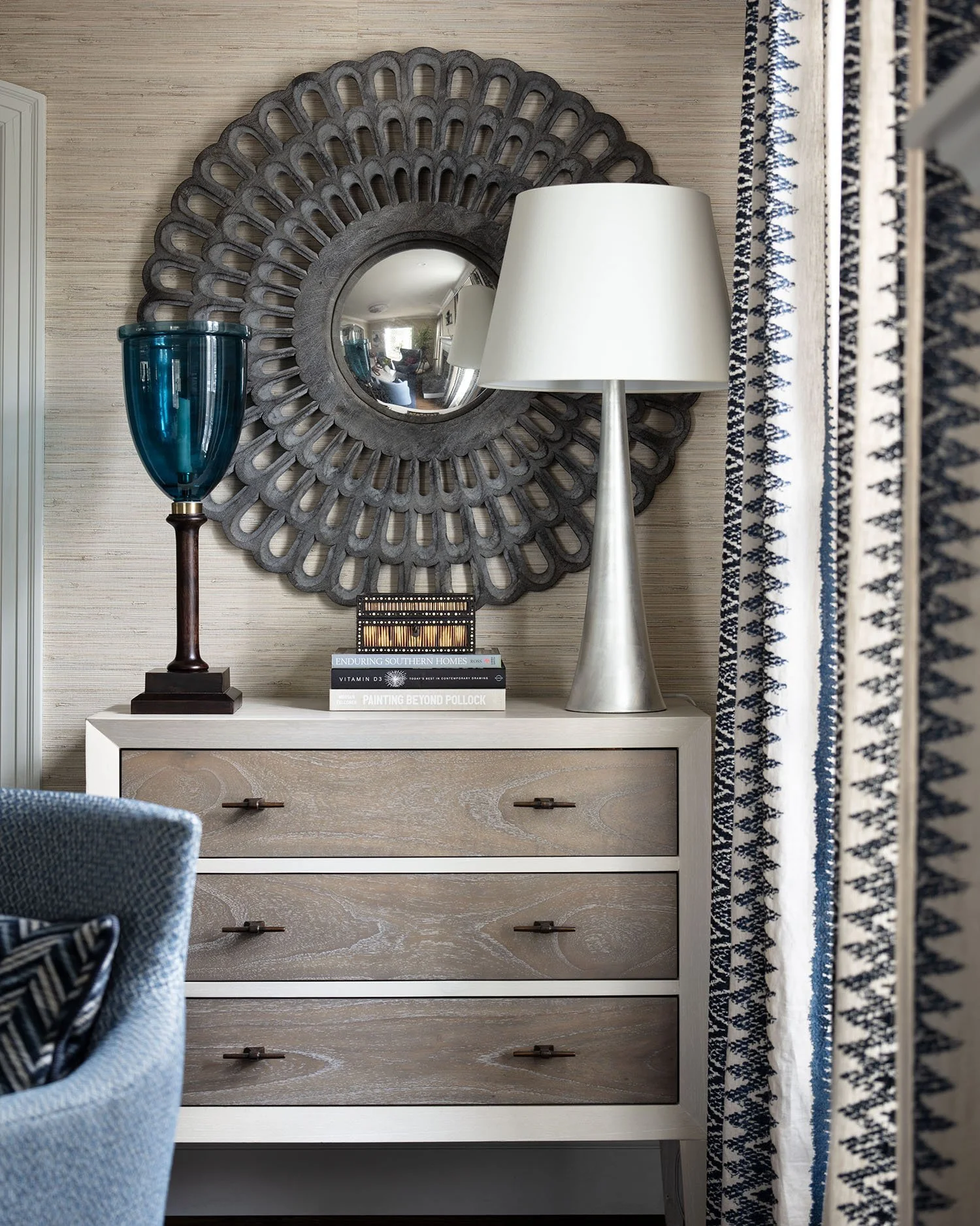 Decorative console table with a large round mirror, a blue glass vase, a white table lamp, and books, set against a textured wall and patterned curtains.