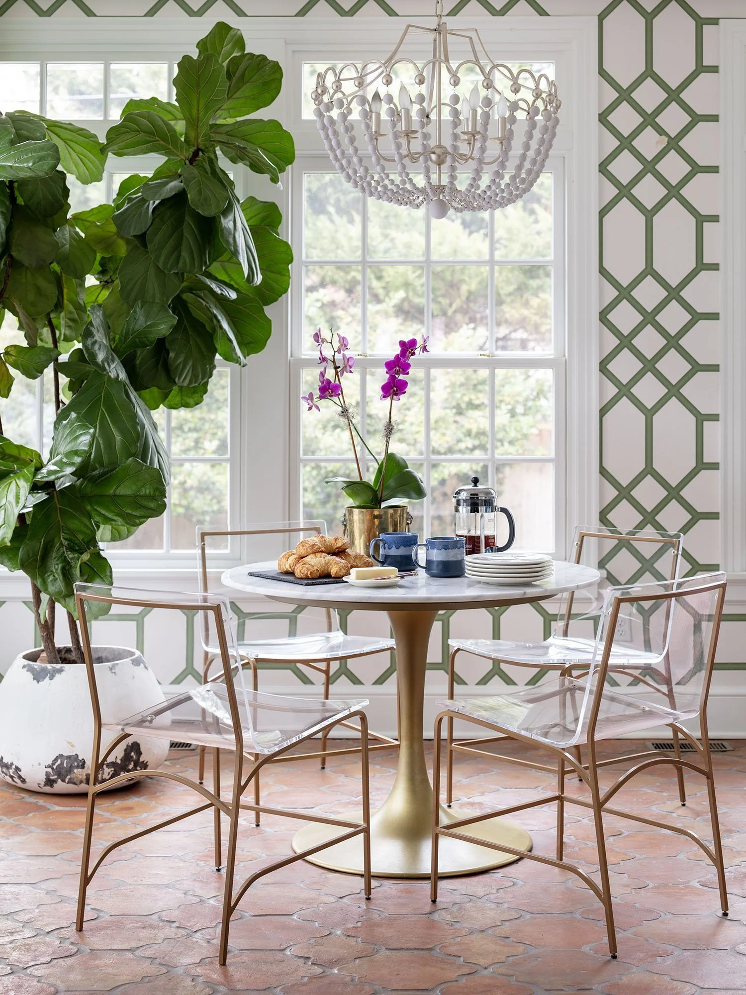 A bright dining area with a round marble table, four clear chairs with metal frames, and a large green potted plant in a white pot. The table is set with croissants, a purple orchid in a gold vase, cups, a French press, and plates. A large window let