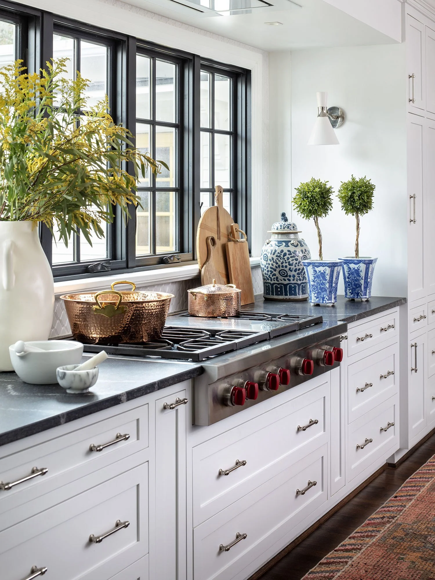 Modern kitchen with white cabinets, dark stone countertop, copper pots, cutting boards, decorative blue and white jars and planters, large window with black framing, and a colorful rug.