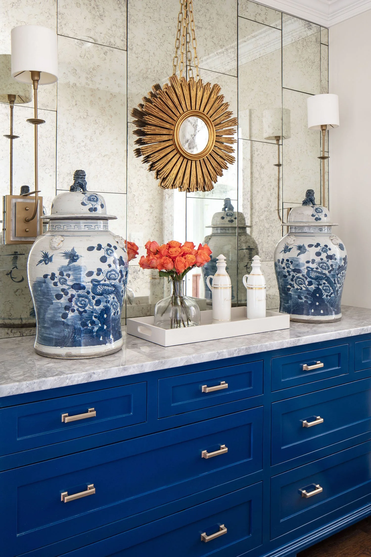 A bathroom sideboard with a marble top, blue drawers with gold handles, two large blue and white porcelain vases, a glass vase with pink roses, and decorative white bottles. Above the sideboard, a mirror is flanked by two wall sconces, and an ornate 