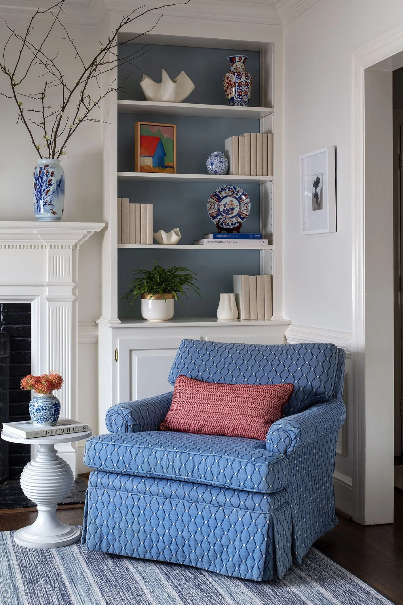 Living room corner featuring a blue patterned armchair with a pink throw pillow, a white side table with a blue vase and orange flowers, a built-in bookshelf with decorative items, a fireplace, and a framed black and white photo on the wall.