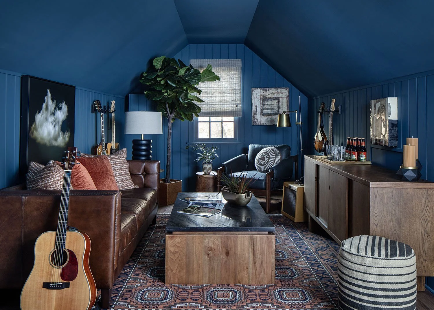 Cozy living room with blue walls, brown leather sofa, acoustic guitar, various artwork, a large potted plant, and a window with woven shade.